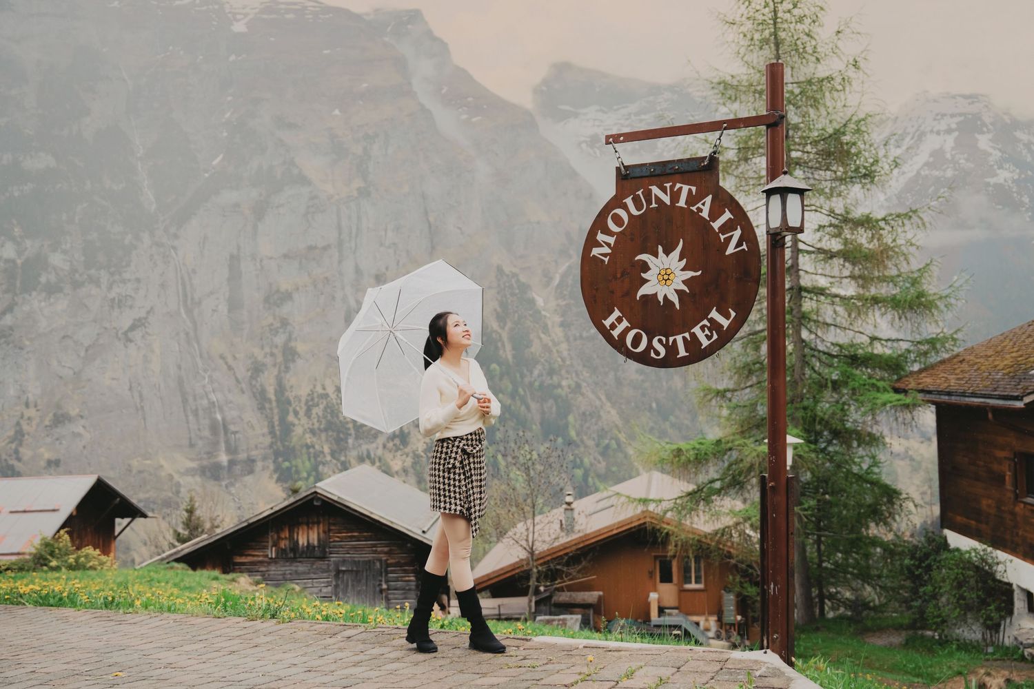 Woman walking past Mountain Hostel sign with snowy peaks and alpine chalets in background.