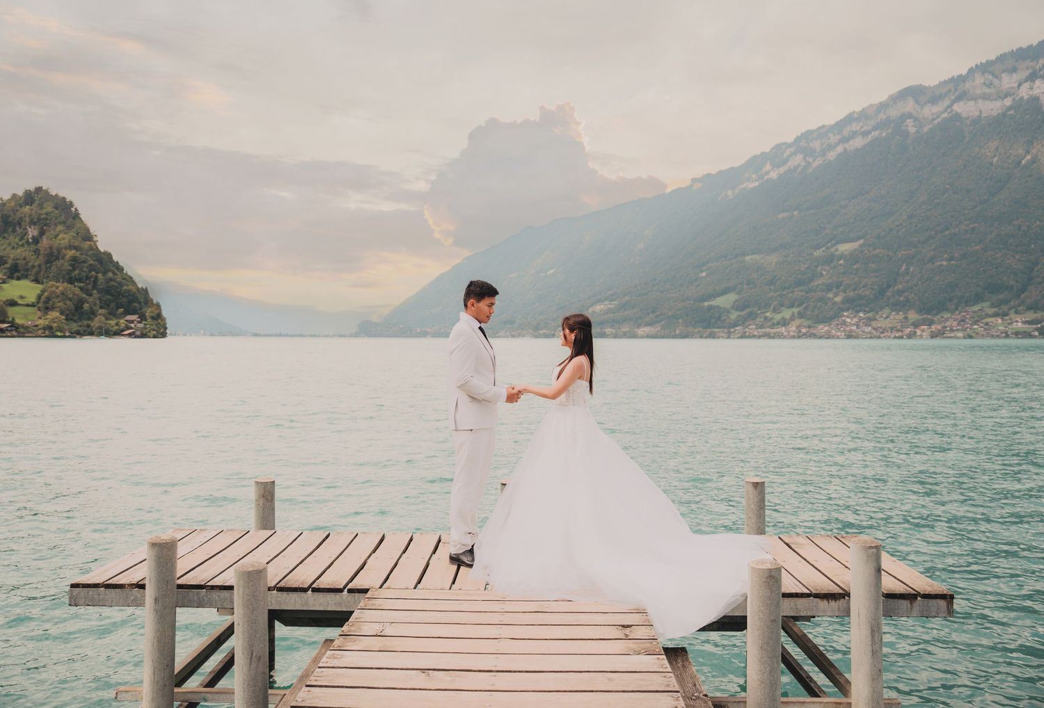 Couple in wedding attire hold hands on a dock surrounded by majestic alpine mountains and turquoise lake.