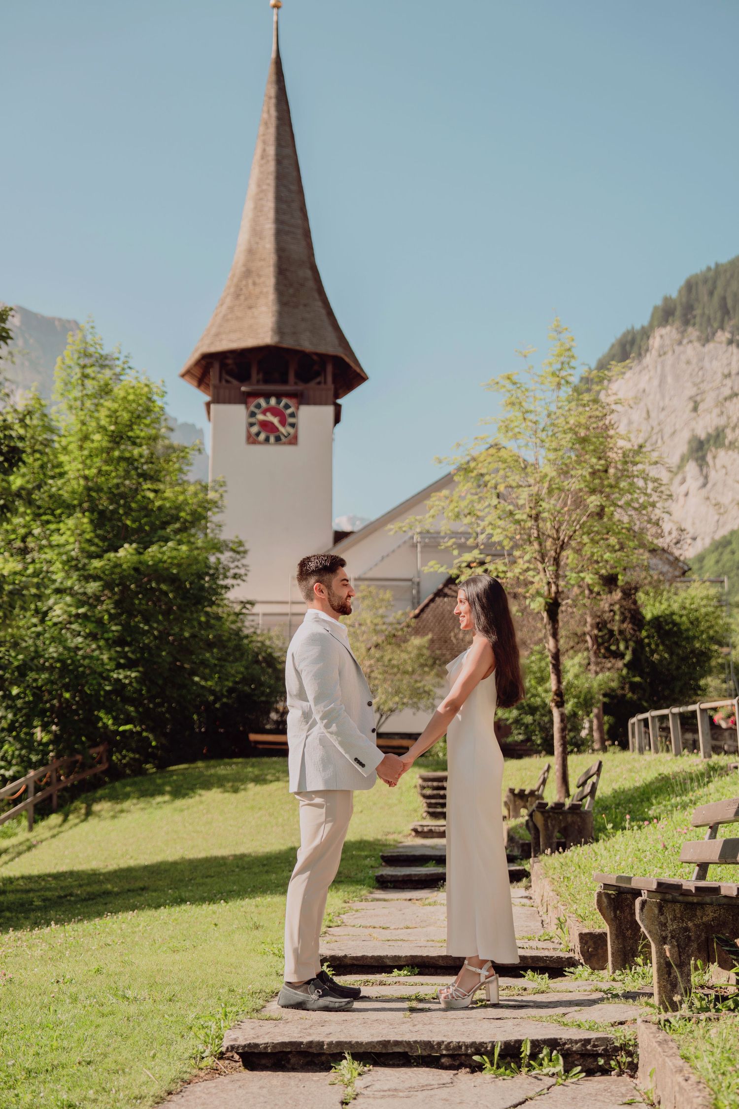 Couple holding hands on a garden path before a charming Swiss church with a pointed steeple and mountain backdrop.