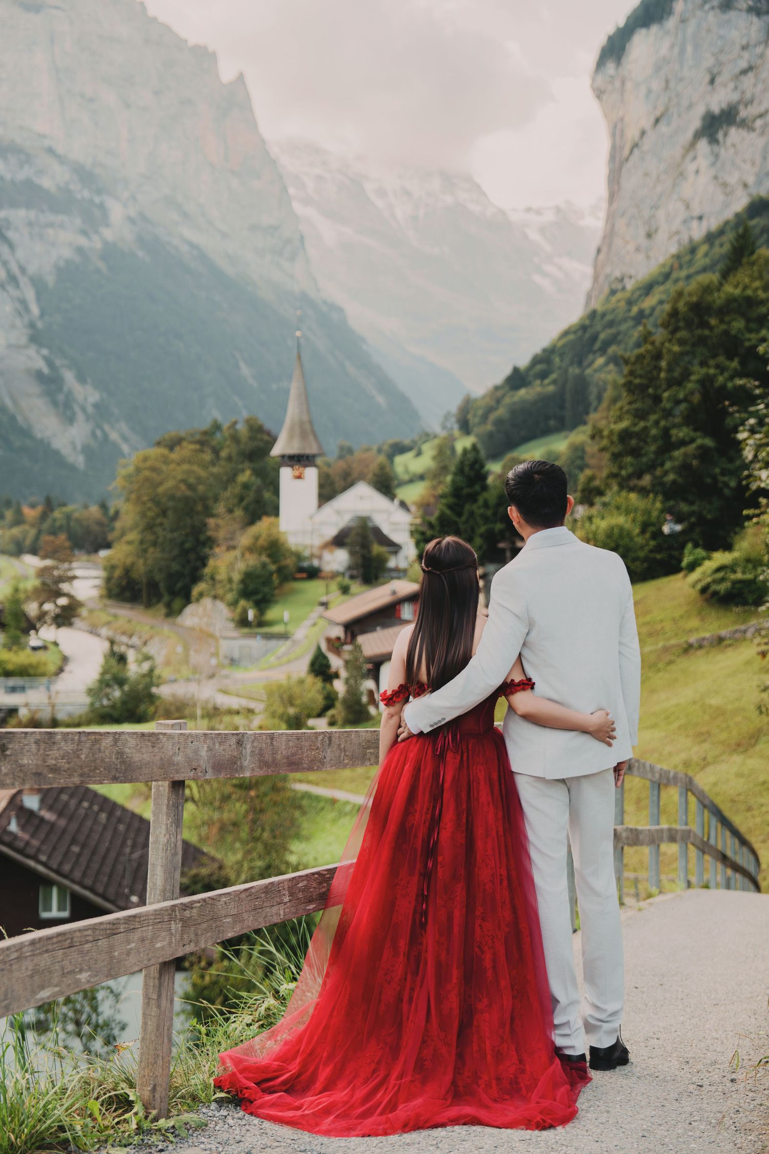 Couple embracing from behind on a bridge overlooking a scenic Swiss alpine village and misty mountains.