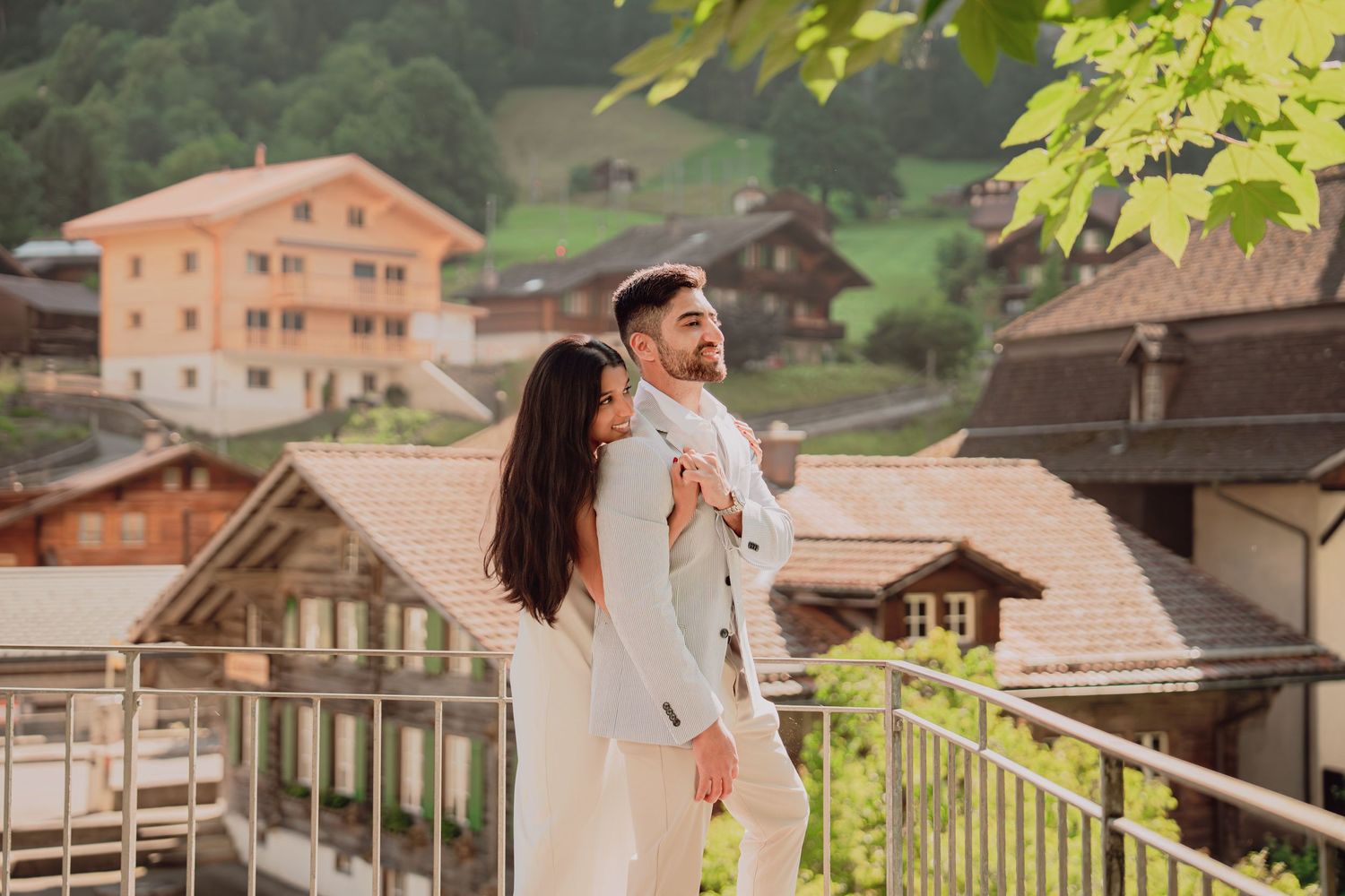 Couple in elegant attire embracing on a balcony with Swiss alpine village backdrop.