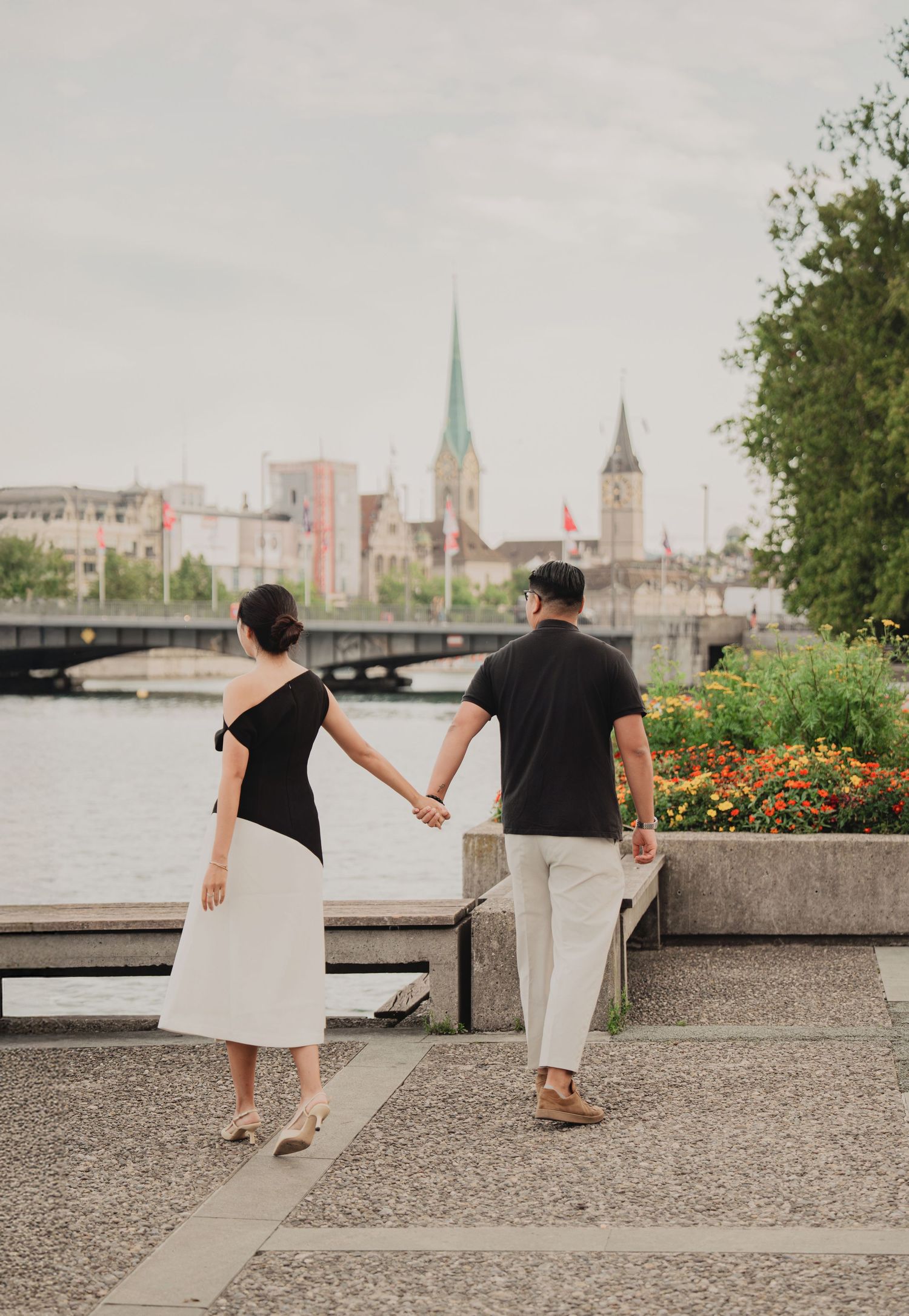 Couple walking hand in hand along Zurich's lakeside promenade with iconic church spires in distance.
