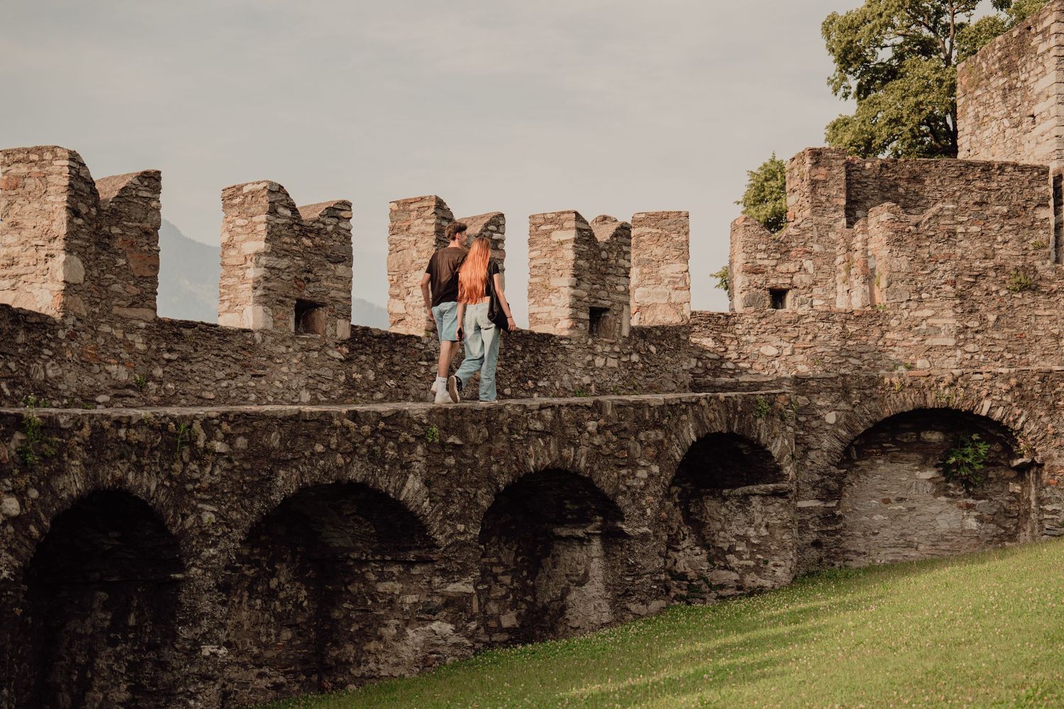 A couple walking along the top of a medieval castle battlement wall surrounded by lush green grounds.