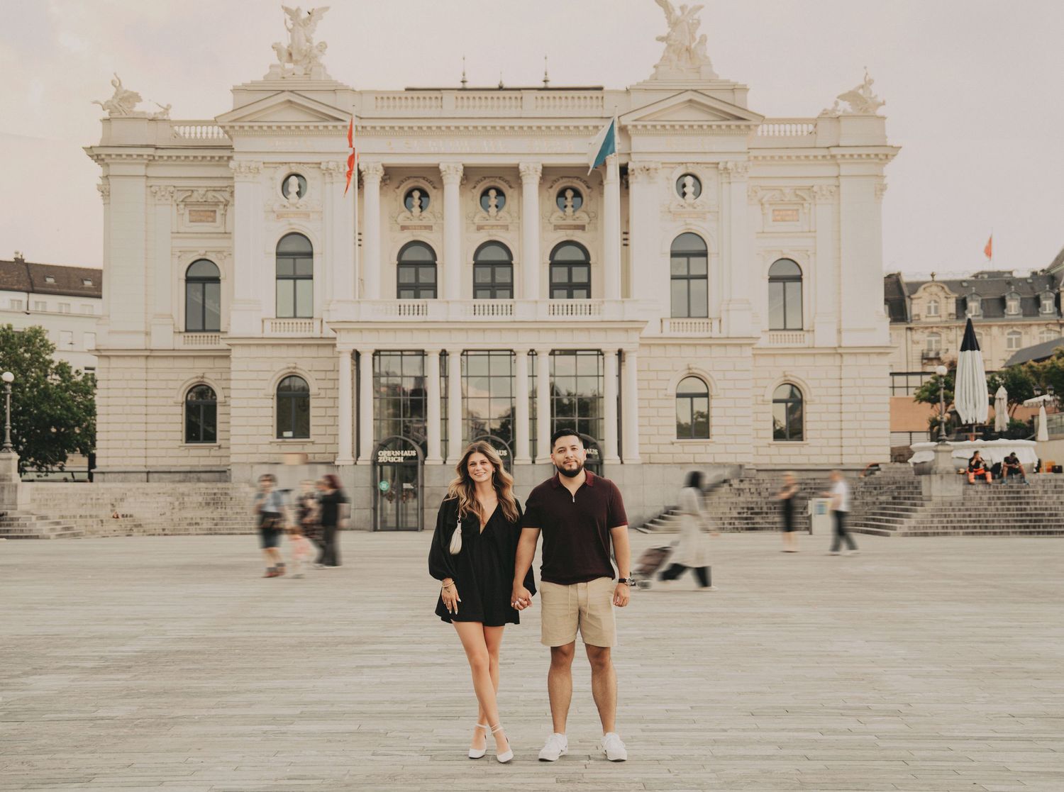 Couple holding hands in front of an ornate white European opera house on a cloudy day.