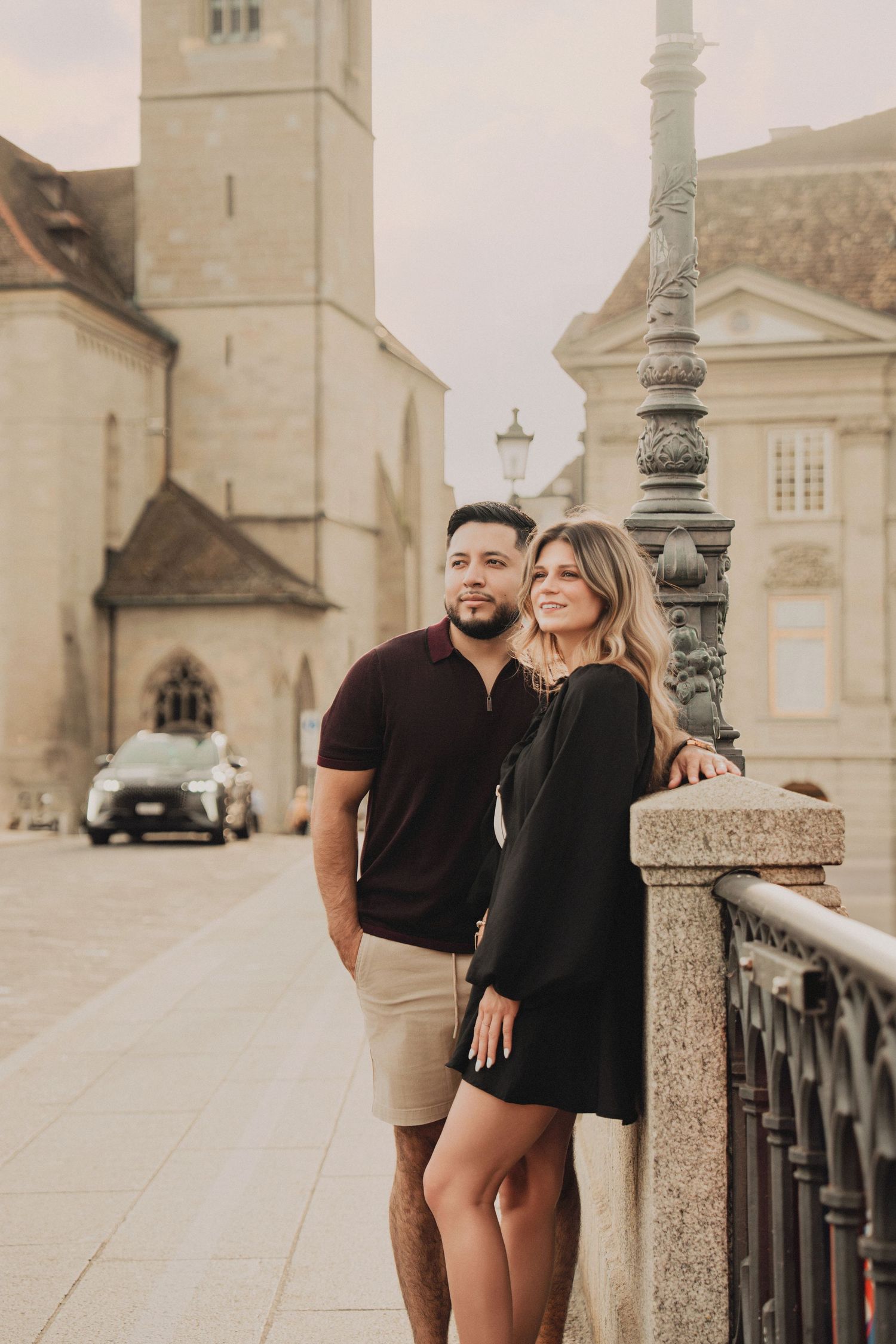 Romantic couple standing by ornate lamppost on Zurich bridge with medieval church behind.