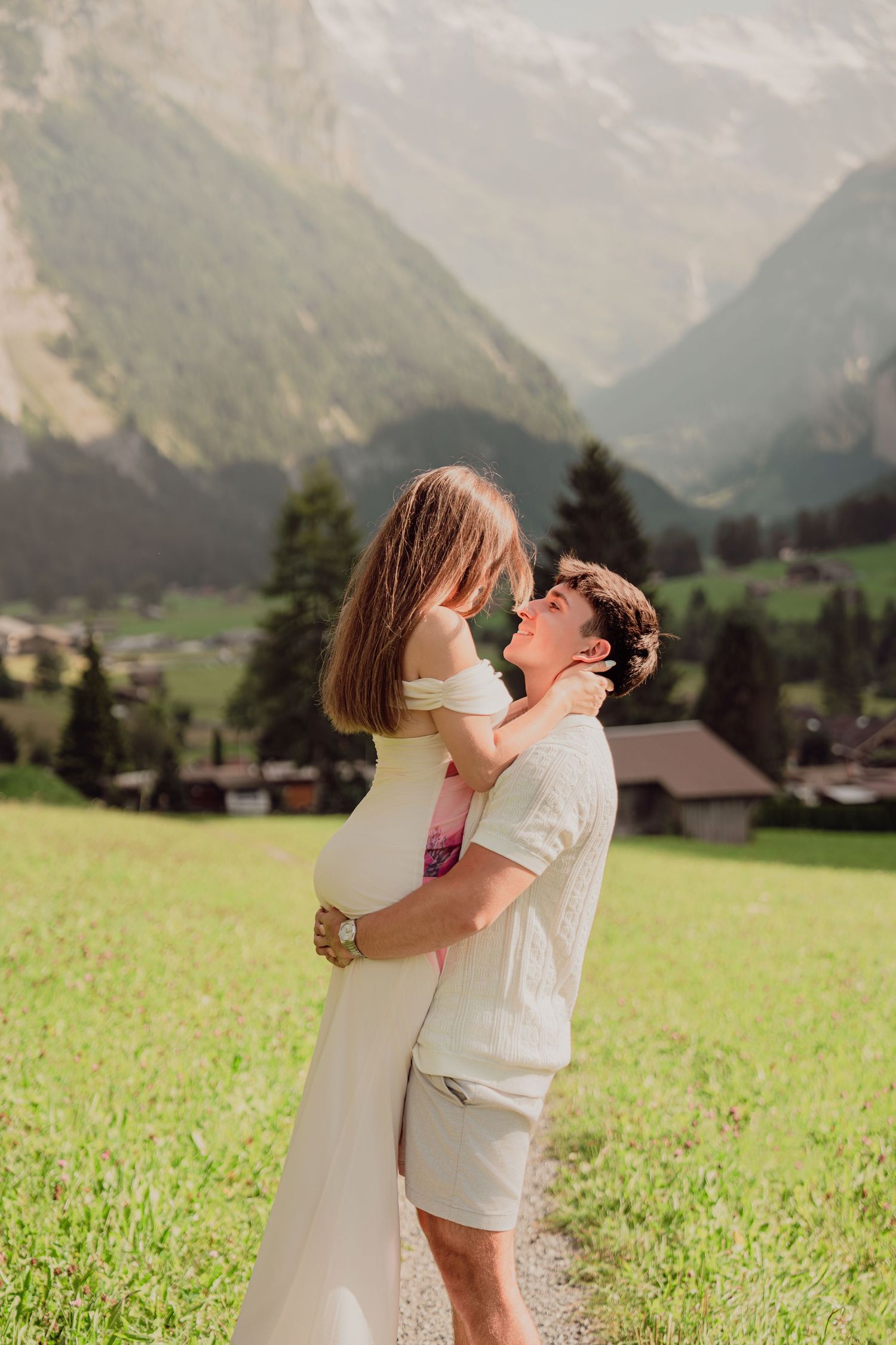 Man lifting woman in white dress on a grassy alpine path with Swiss mountain valley backdrop.