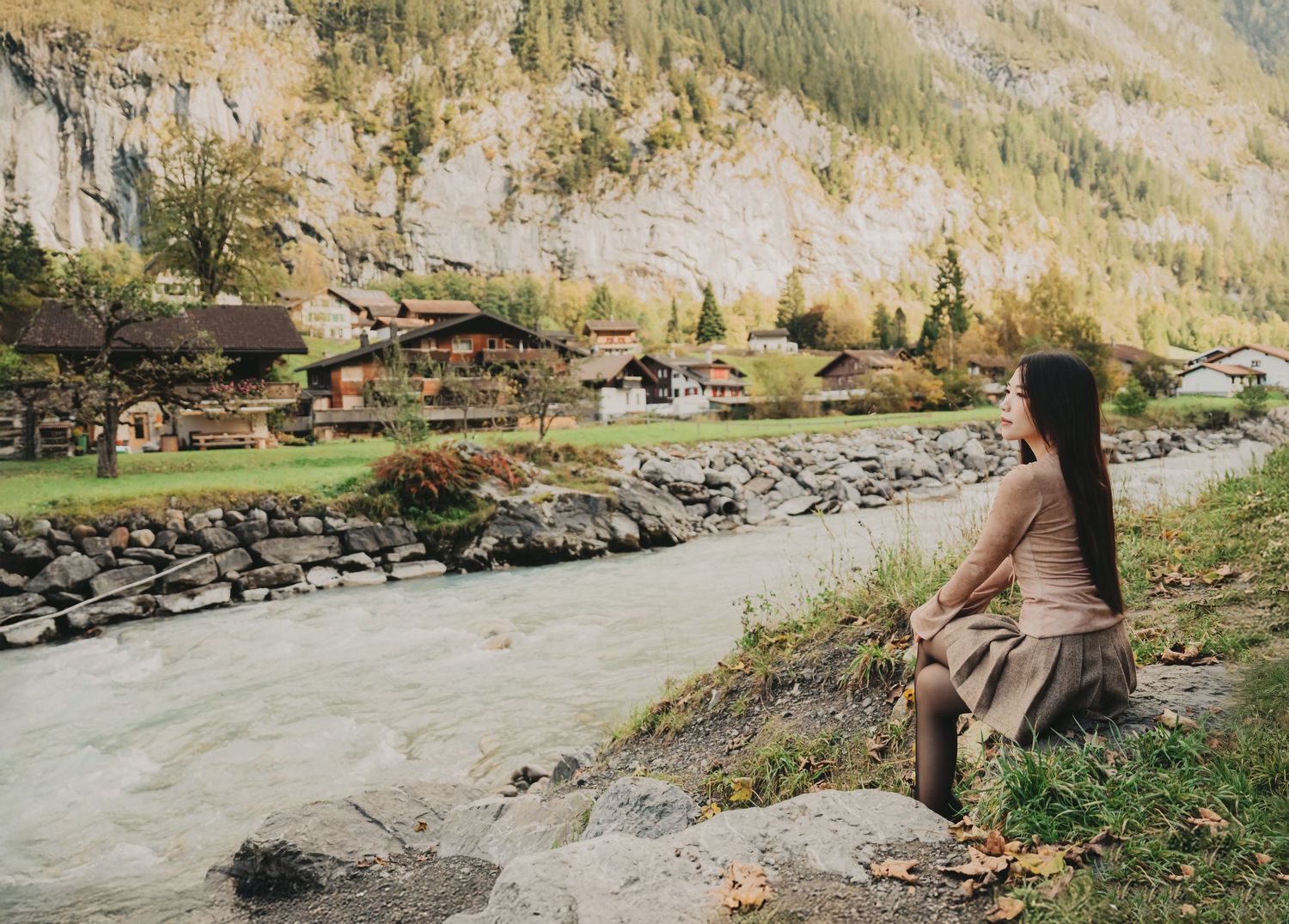Woman sitting on a rock beside a rushing glacial river in a picturesque Swiss alpine valley.