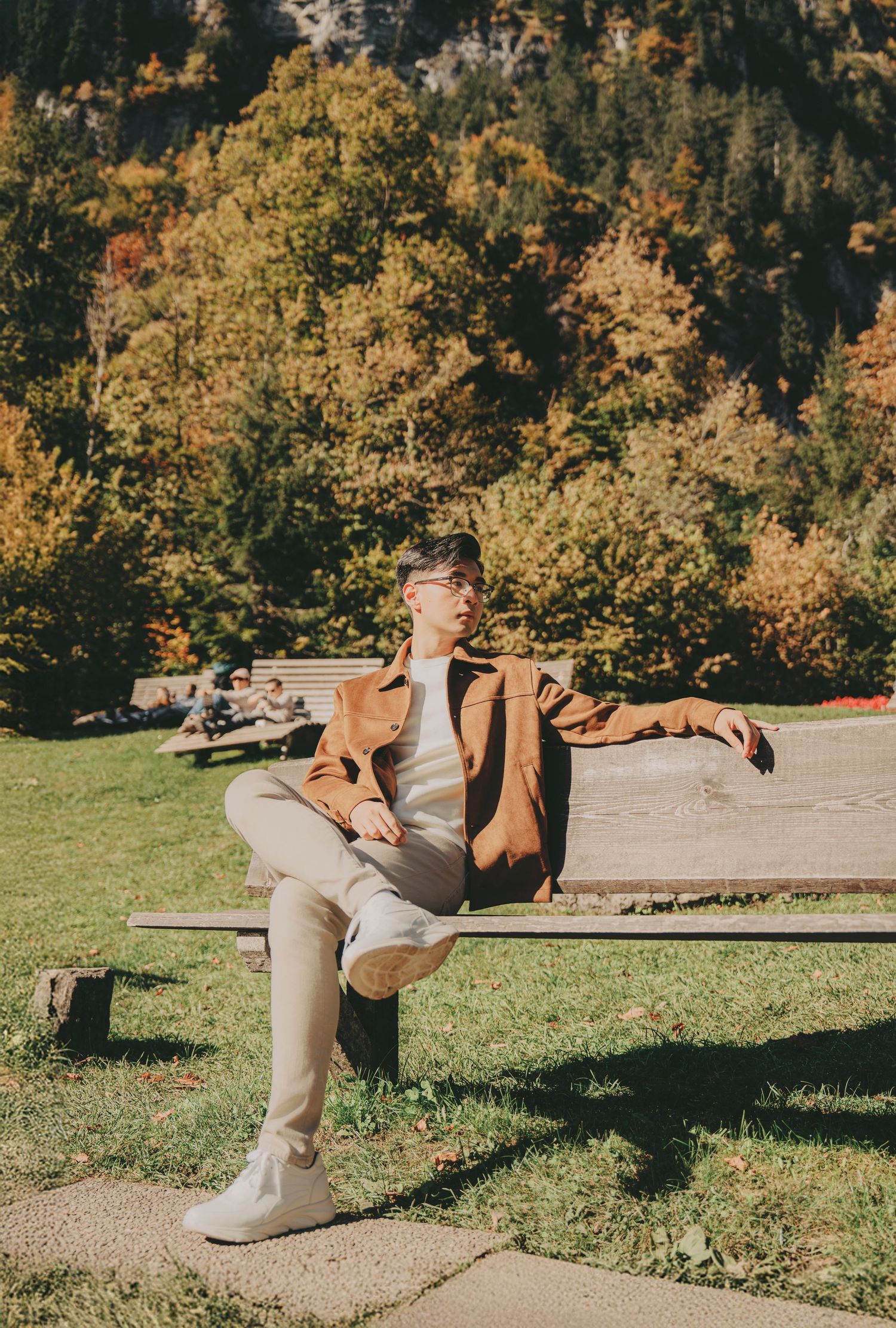 Young man in brown jacket relaxing on wooden bench in sunny autumn park setting.