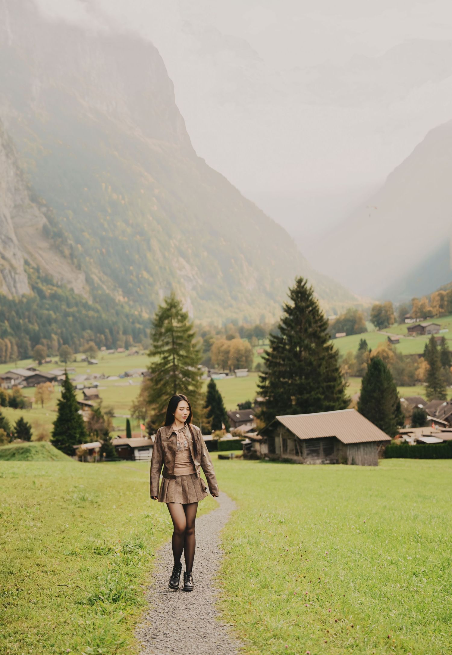Woman in brown outfit strolling on a path through a stunning Swiss alpine valley with tall pines.