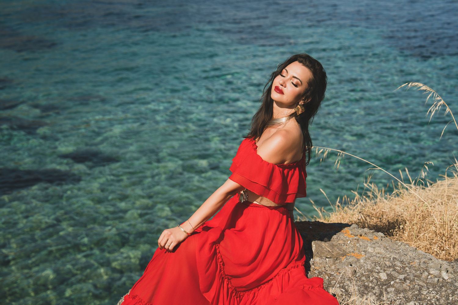 Elegant woman in red off-shoulder dress sitting on rocks by crystal clear turquoise sea water.
