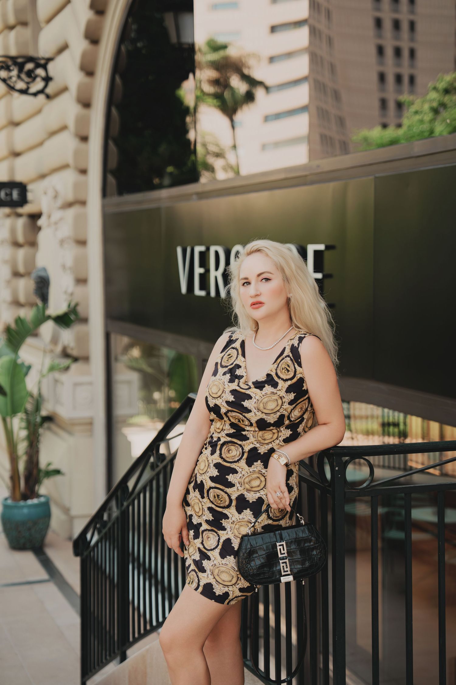 Blonde woman in black and gold Versace patterned dress leaning on railing outside boutique.