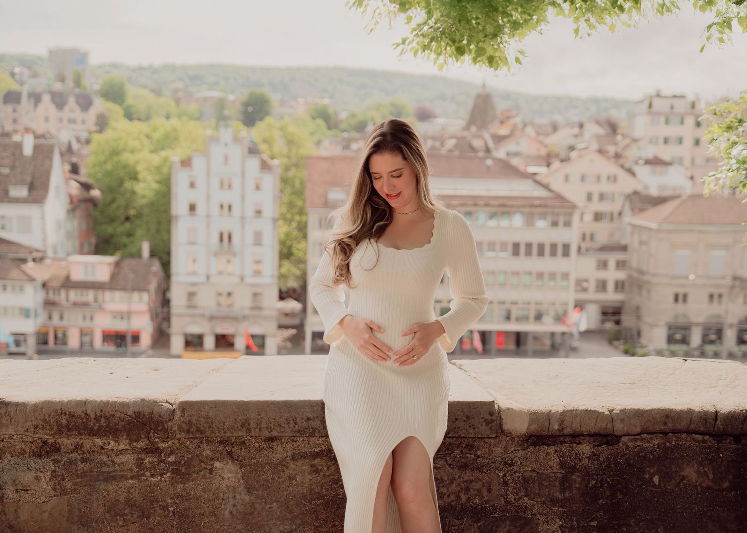 Pregnant woman in white ribbed dress looks down at baby bump against blurred European city backdrop.