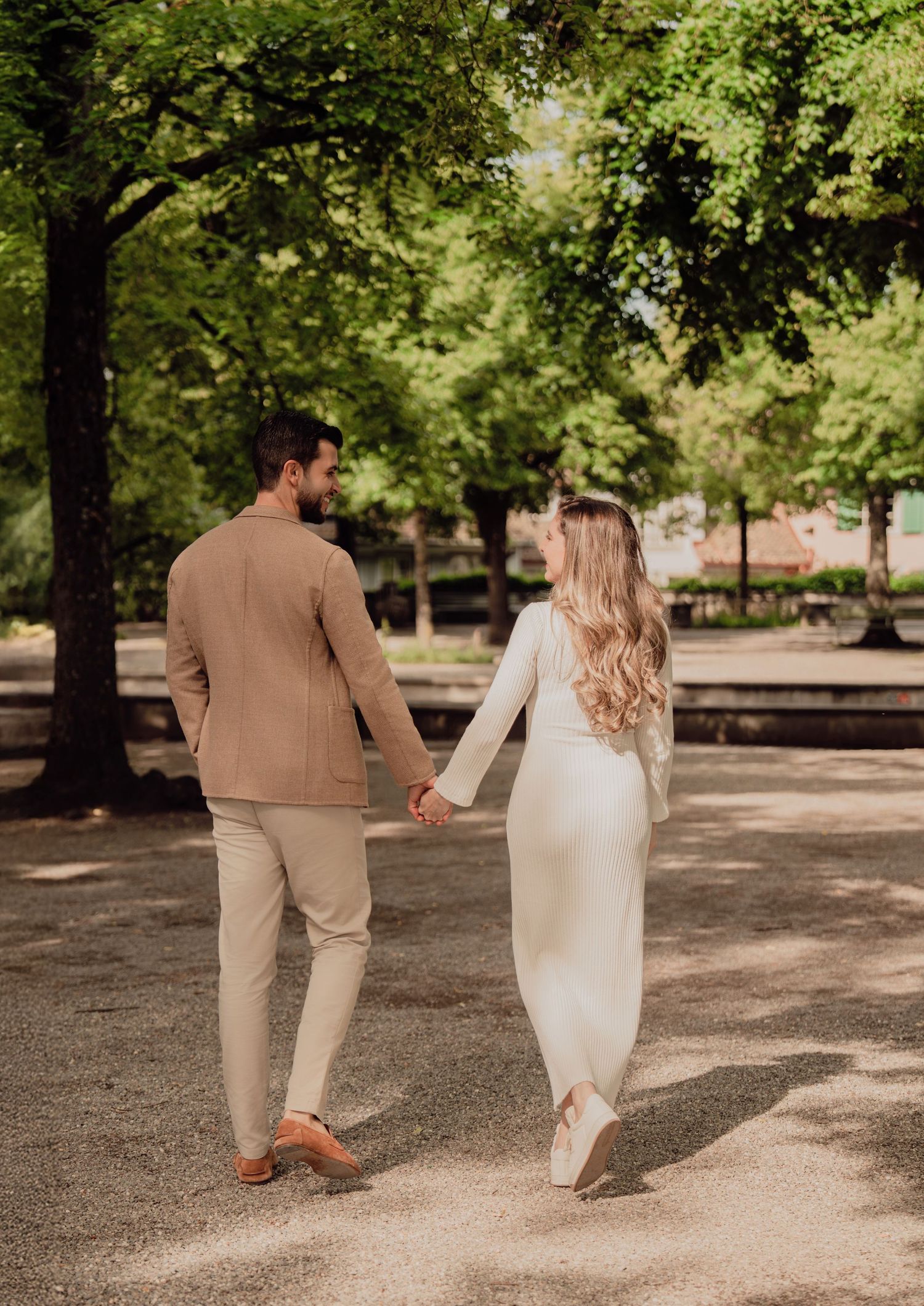 Couple walking away hand in hand through a sunlit park, woman in white dress with long blonde hair, man in tan blazer.