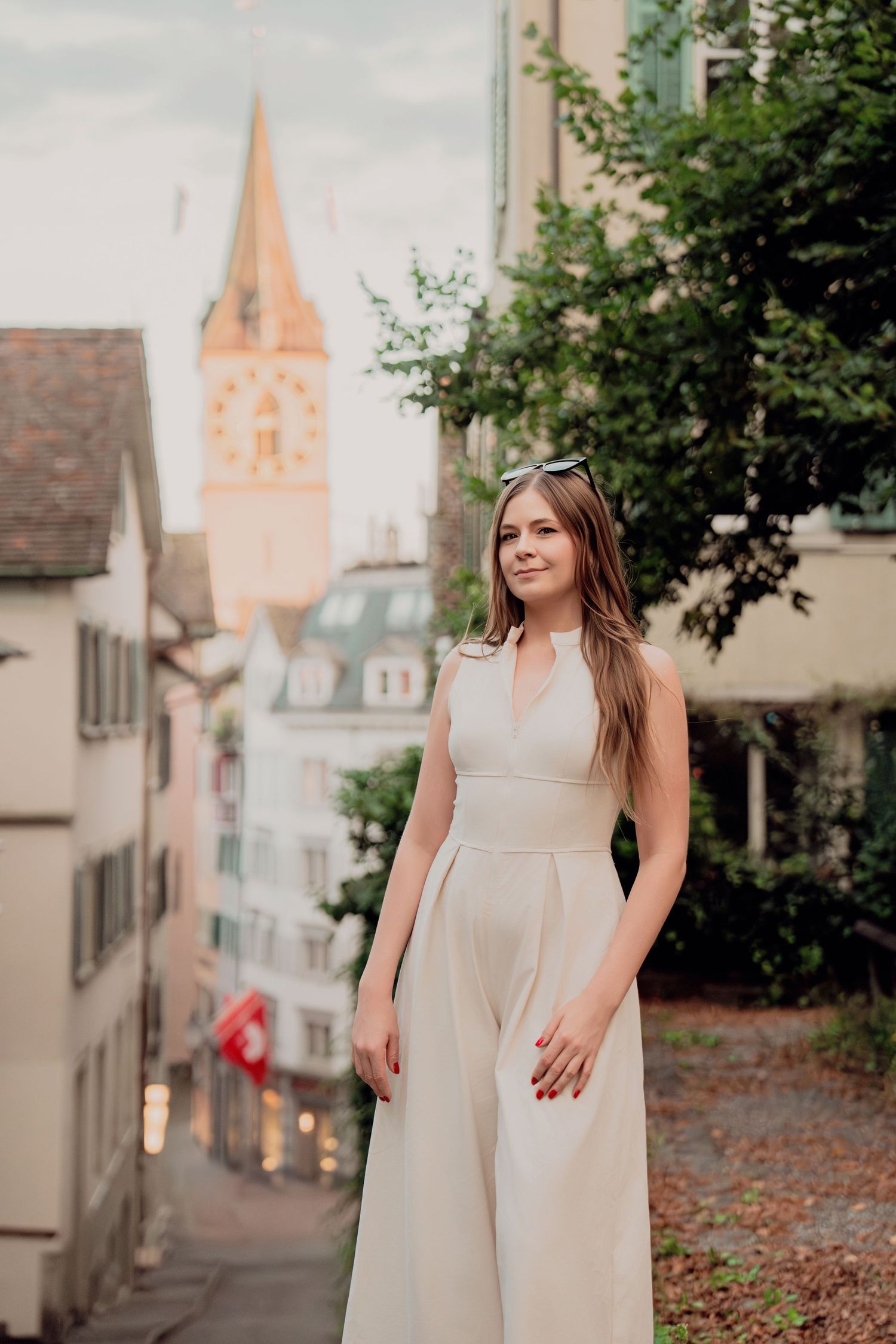 Woman in white jumpsuit smiling on scenic European cobblestone street with medieval clock tower behind her.