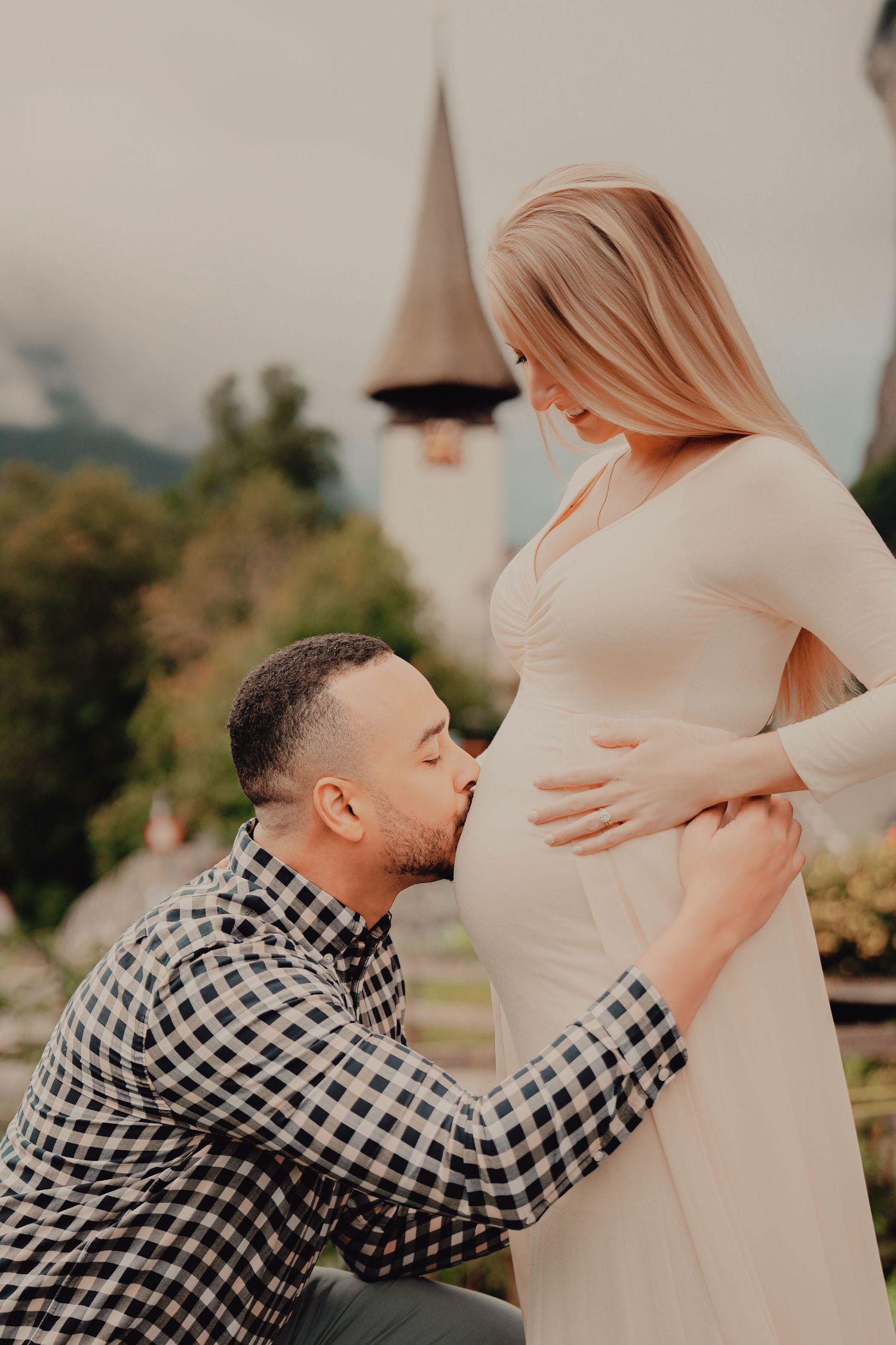 Man kneels to kiss pregnant womans belly outdoors near a church steeple in a scenic mountain village.