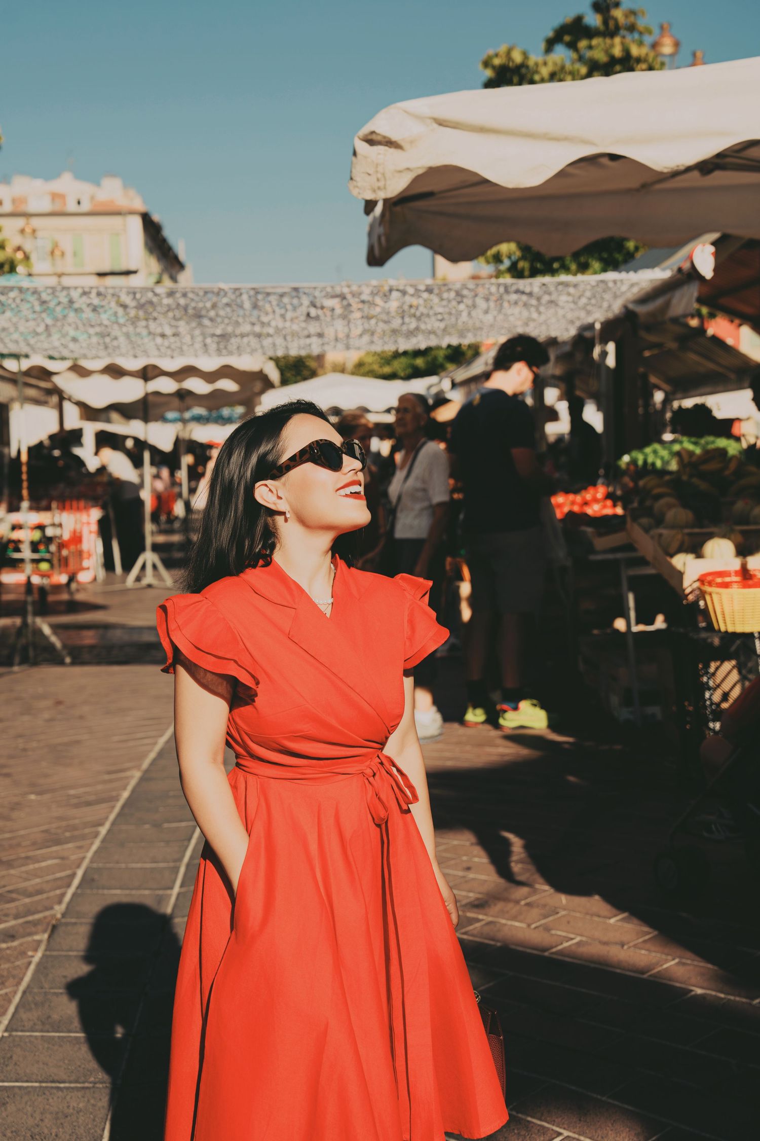 Woman in red wrap dress and sunglasses enjoying sunny outdoor market in city square.