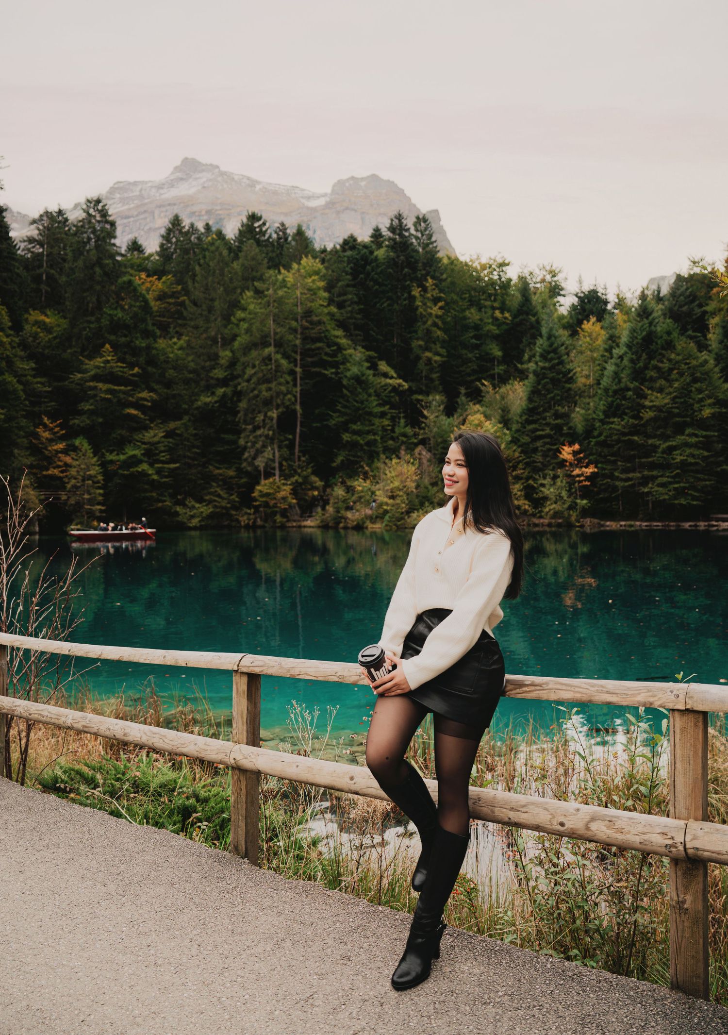 Smiling woman in white sweater black skirt and tall boots posing by stunning turquoise alpine lake.