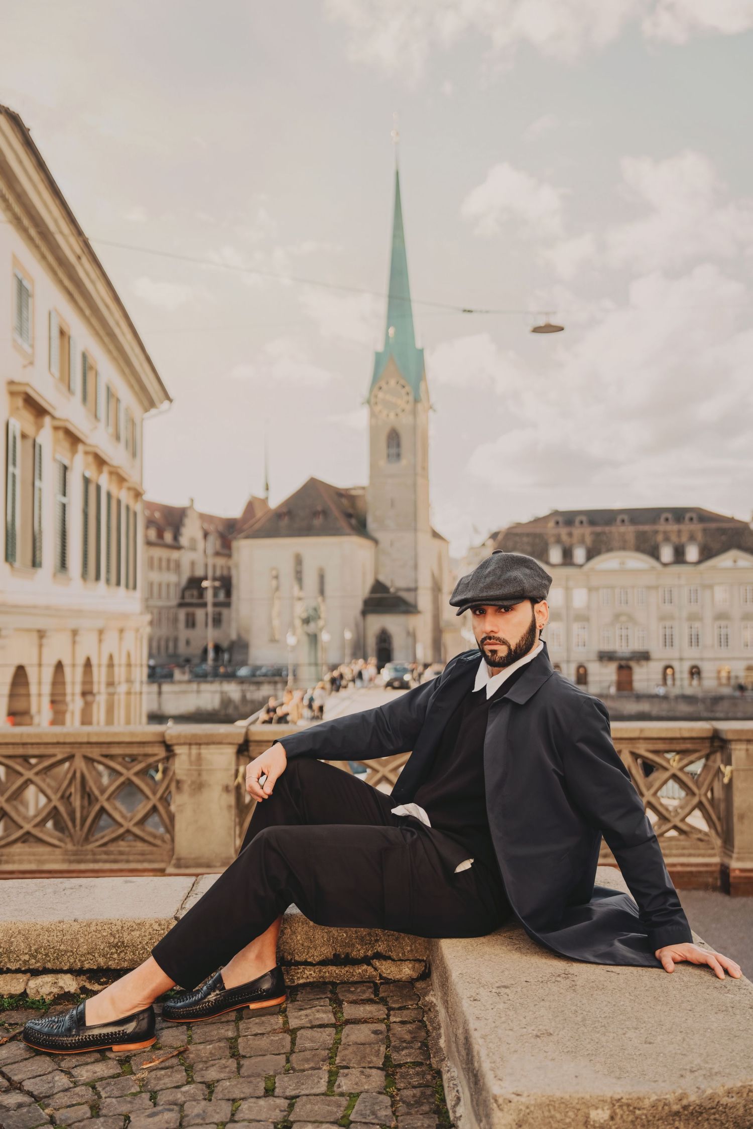 Well-dressed bearded man in dark coat and newsboy cap sits on stone ledge in historic Zurich city square.