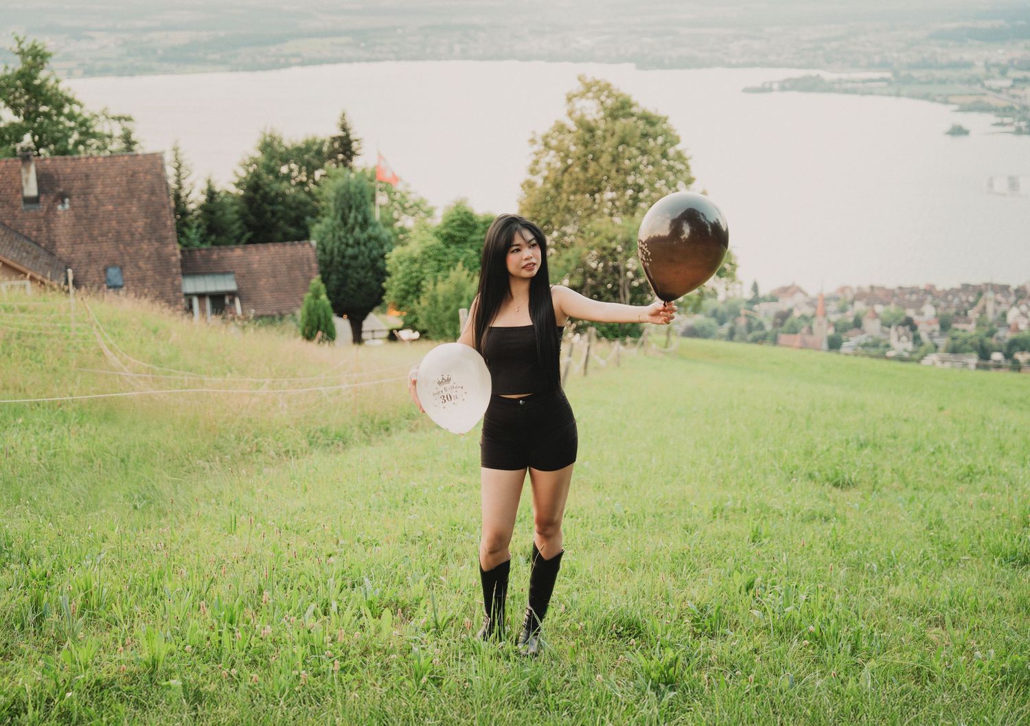 Young woman in black romper and knee-high boots holding metallic balloons on a green hillside with lake view.