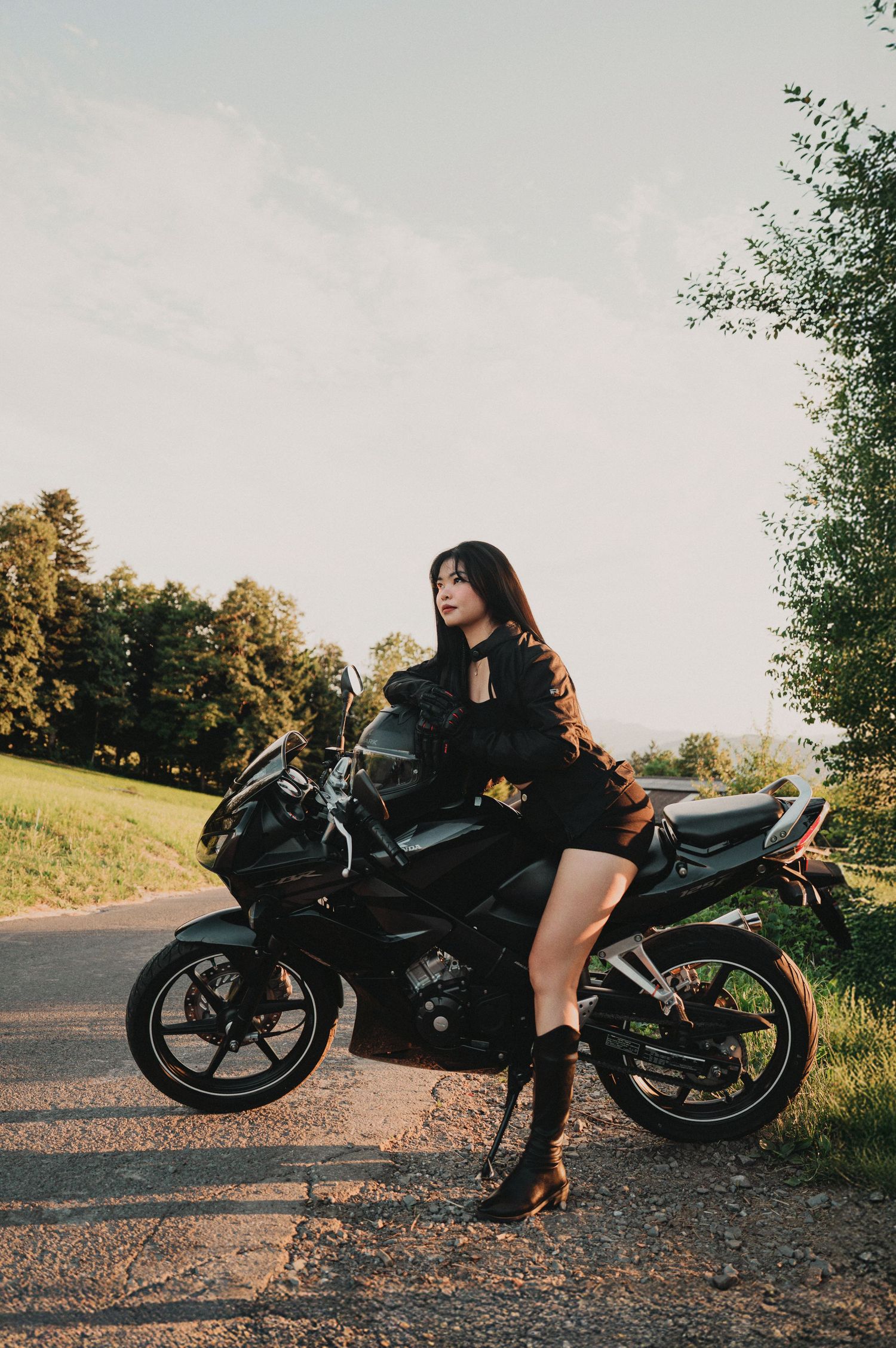 Woman in black outfit leaning against a black sport bike on a rural road with green fields at sunset.