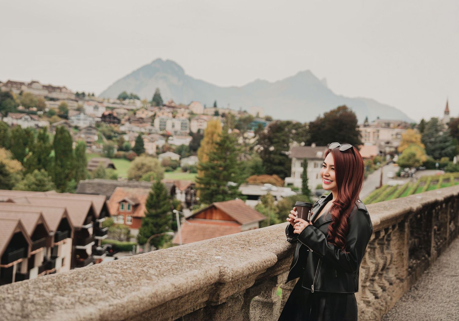 Smiling red-haired woman in leather jacket on stone bridge overlooking a scenic European mountain village.