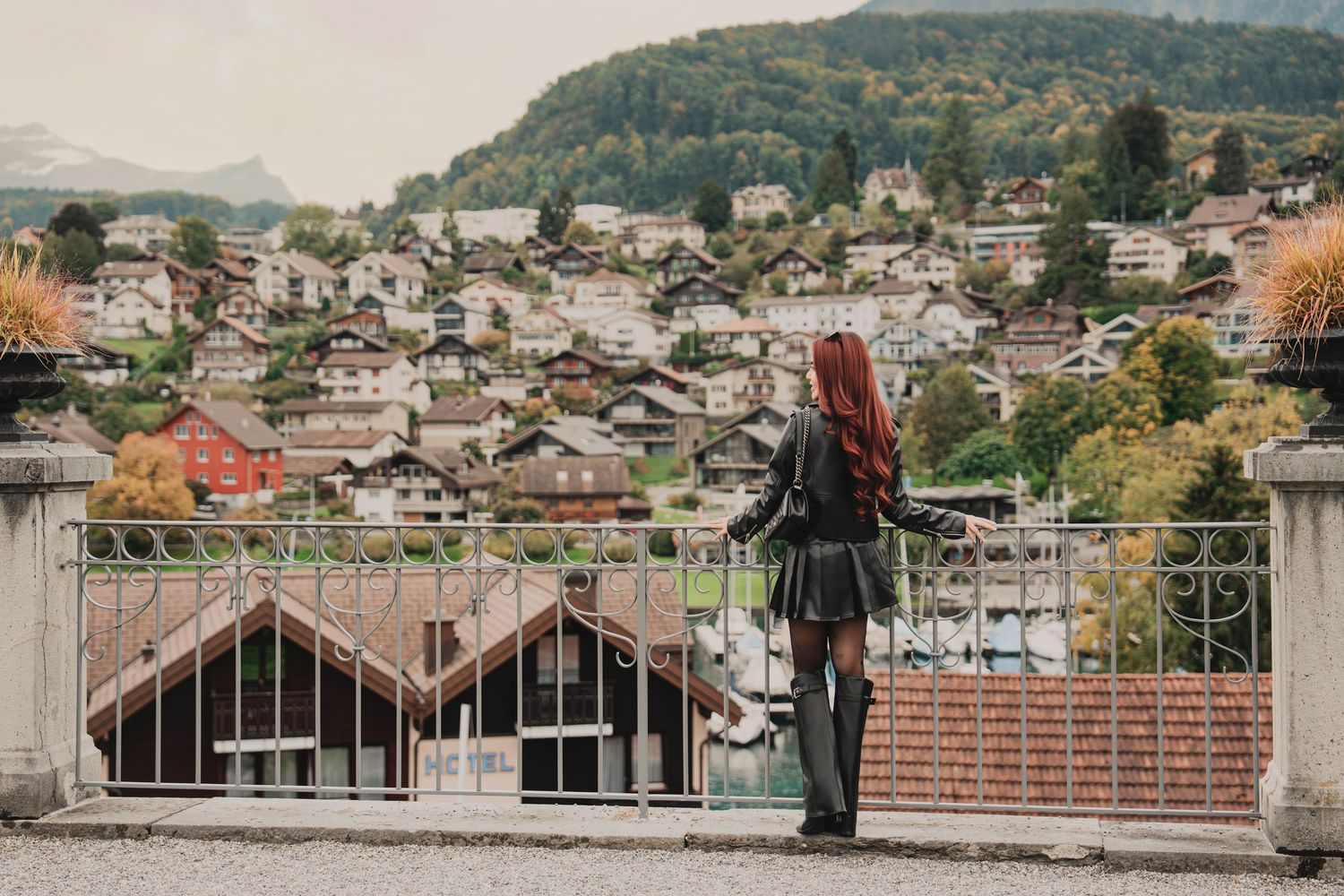 Red-haired woman in black leather outfit stands at railing overlooking scenic Swiss alpine village.