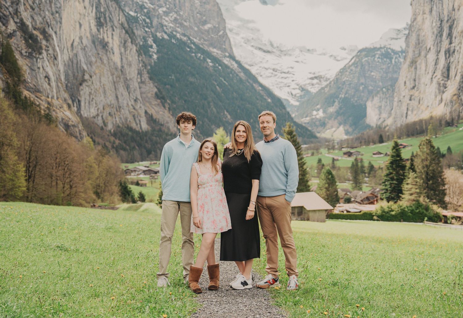 Family of four posing together in Swiss alpine valley with dramatic mountain backdrop and green meadows.