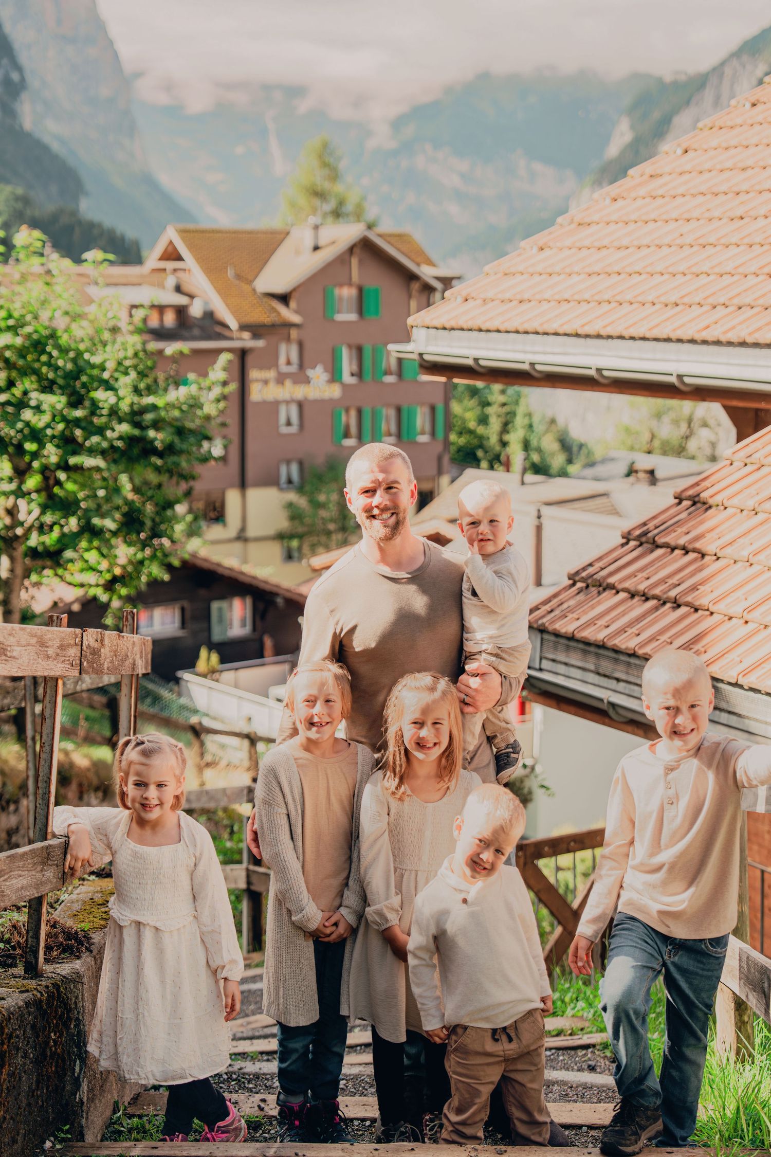 Smiling bald father posing with seven blonde children on steps in a scenic Swiss mountain village.