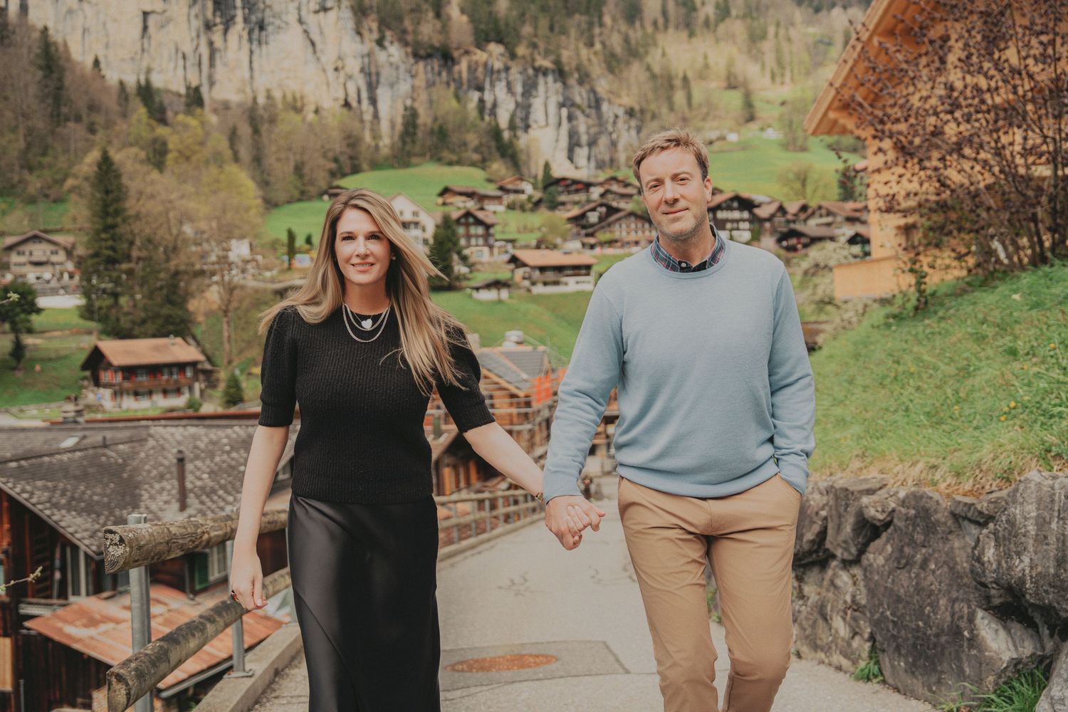 Smiling couple holding hands on a road with scenic Swiss alpine village and cliffs in background.