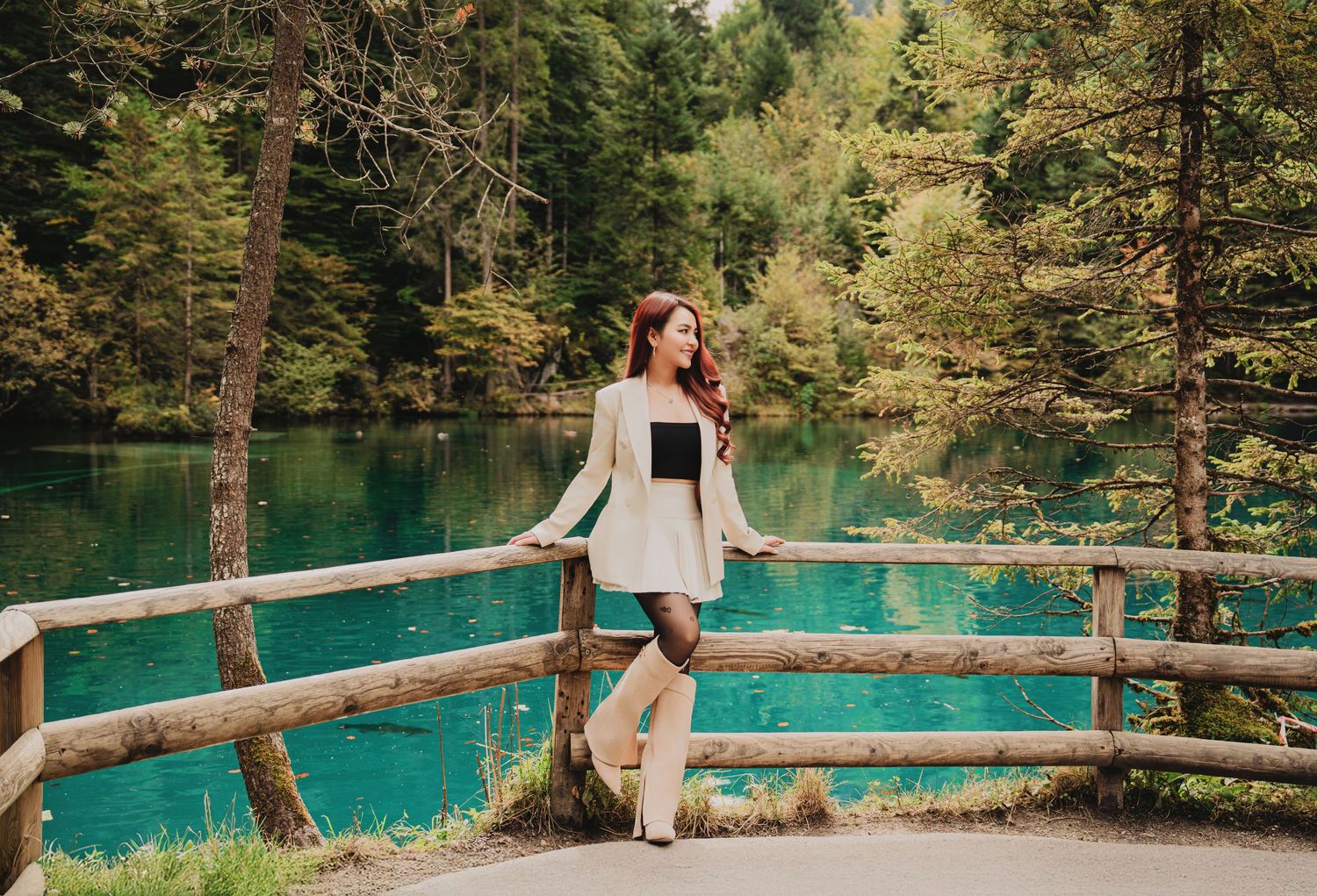 Stylish woman in white blazer leaning on wooden fence by vibrant turquoise forest lake.