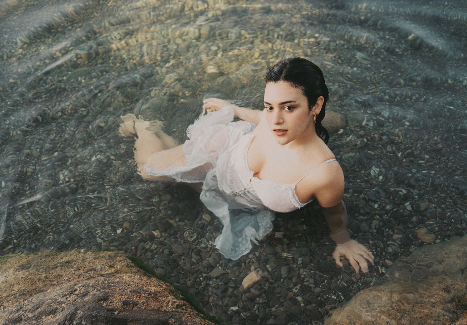 Young woman in sheer white dress floating gracefully in clear shallow water over rocky riverbed.