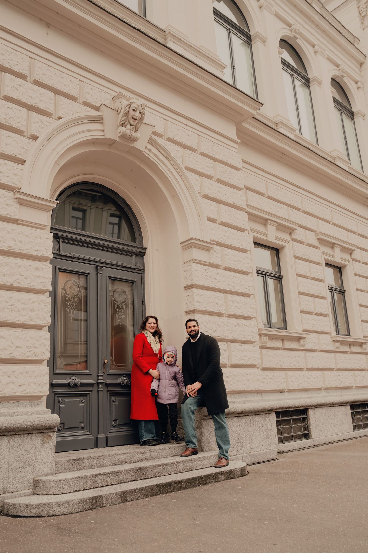 Family of three standing on steps before an elegant arched doorway of a historic building.