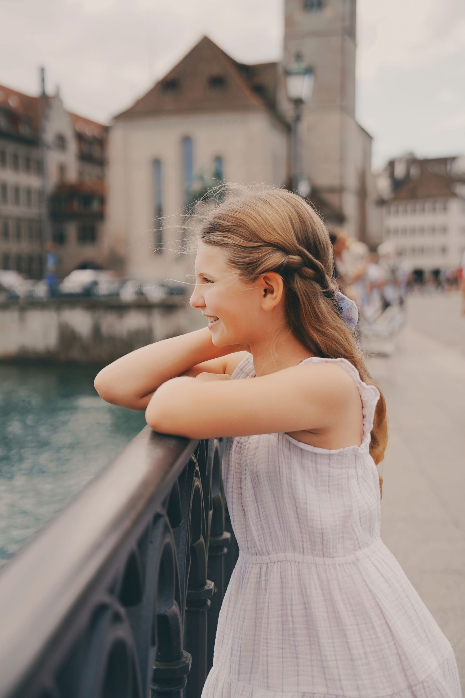 A young blonde girl leans on a bridge railing smiling, with Zurich old town buildings and river in the background.