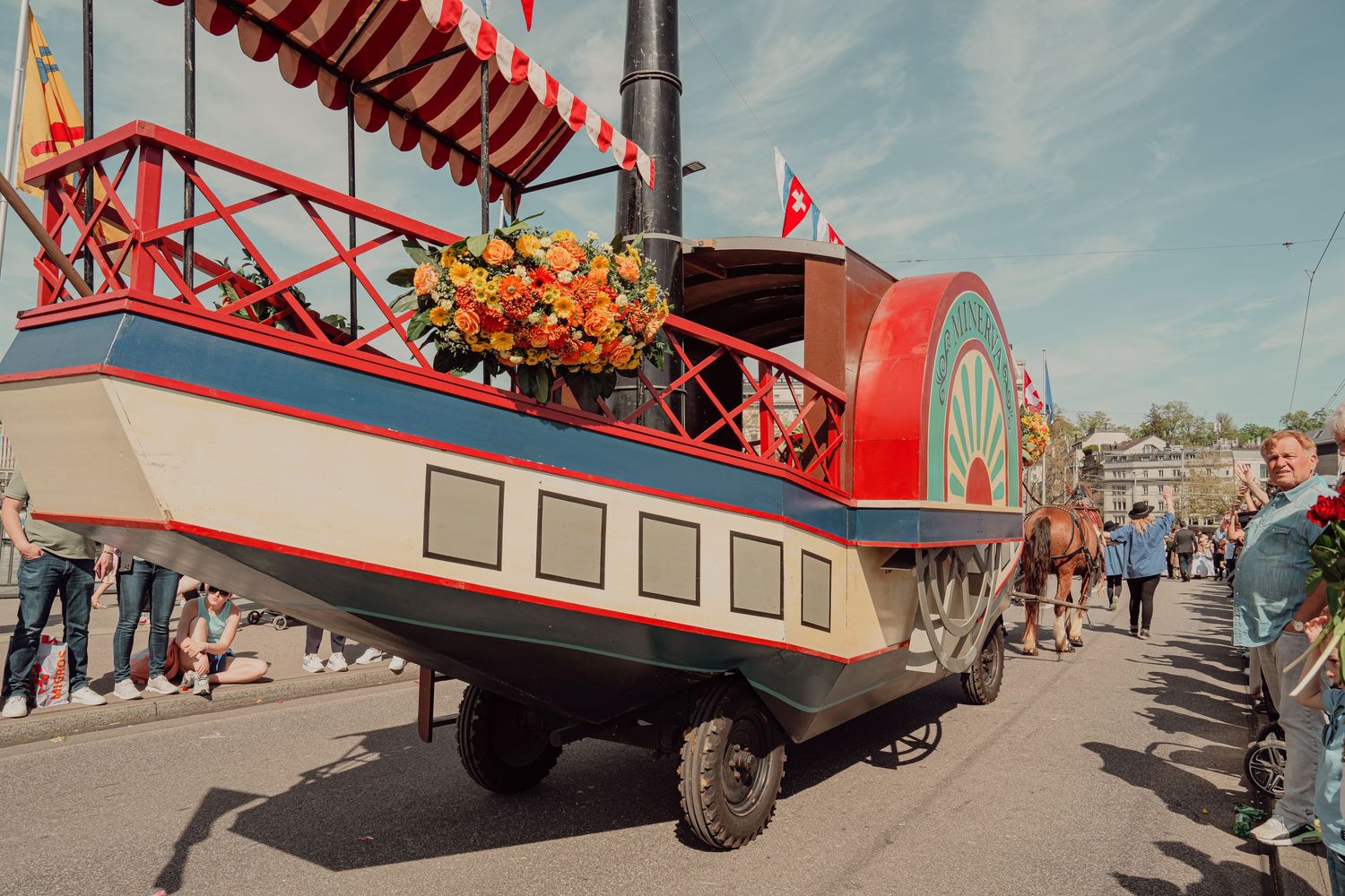 Vintage steamboat parade float decorated with orange flowers and red striped canopy moving through a Swiss festival.