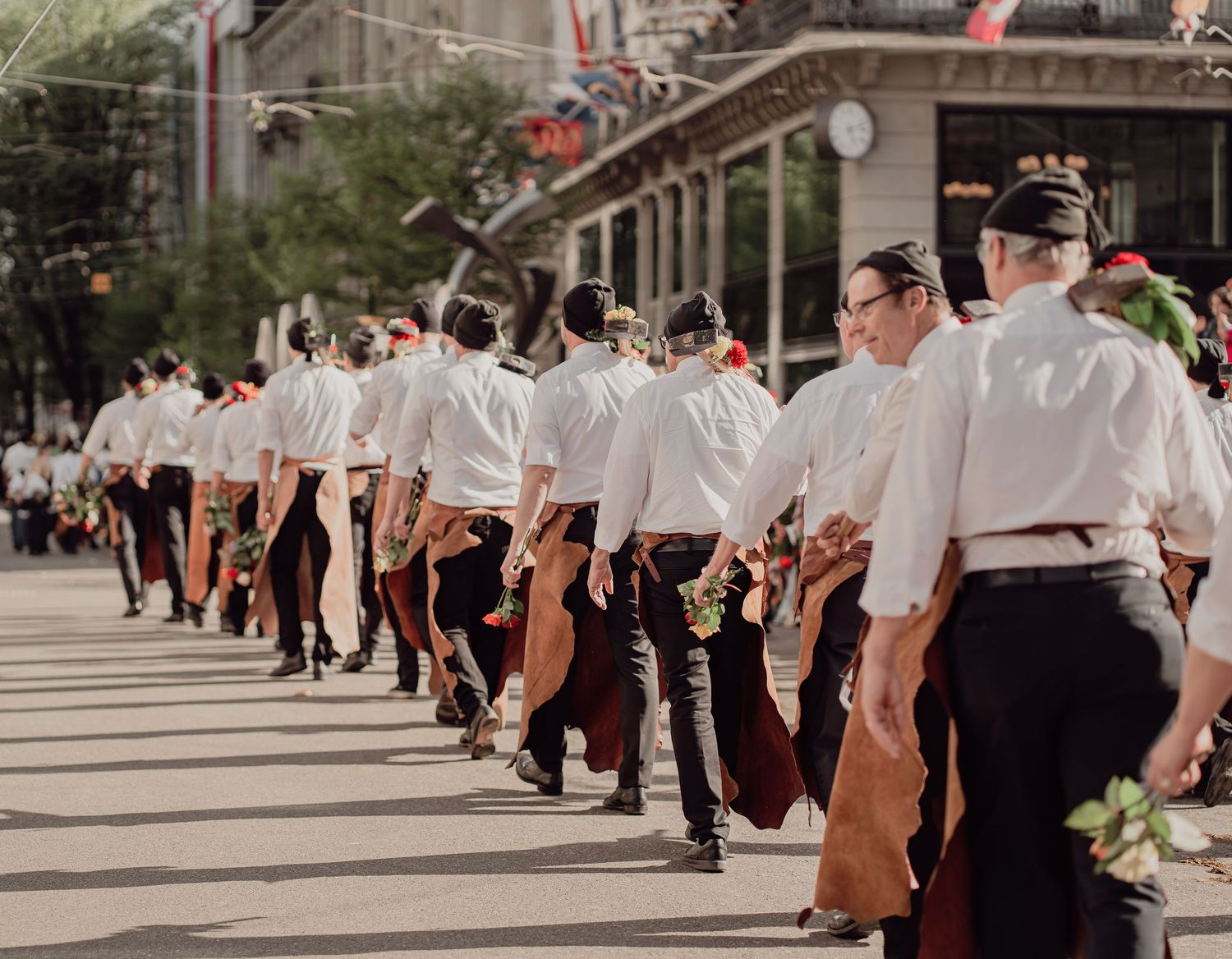 Men in traditional folk costumes march in a parade carrying flowers along a sunny city street.