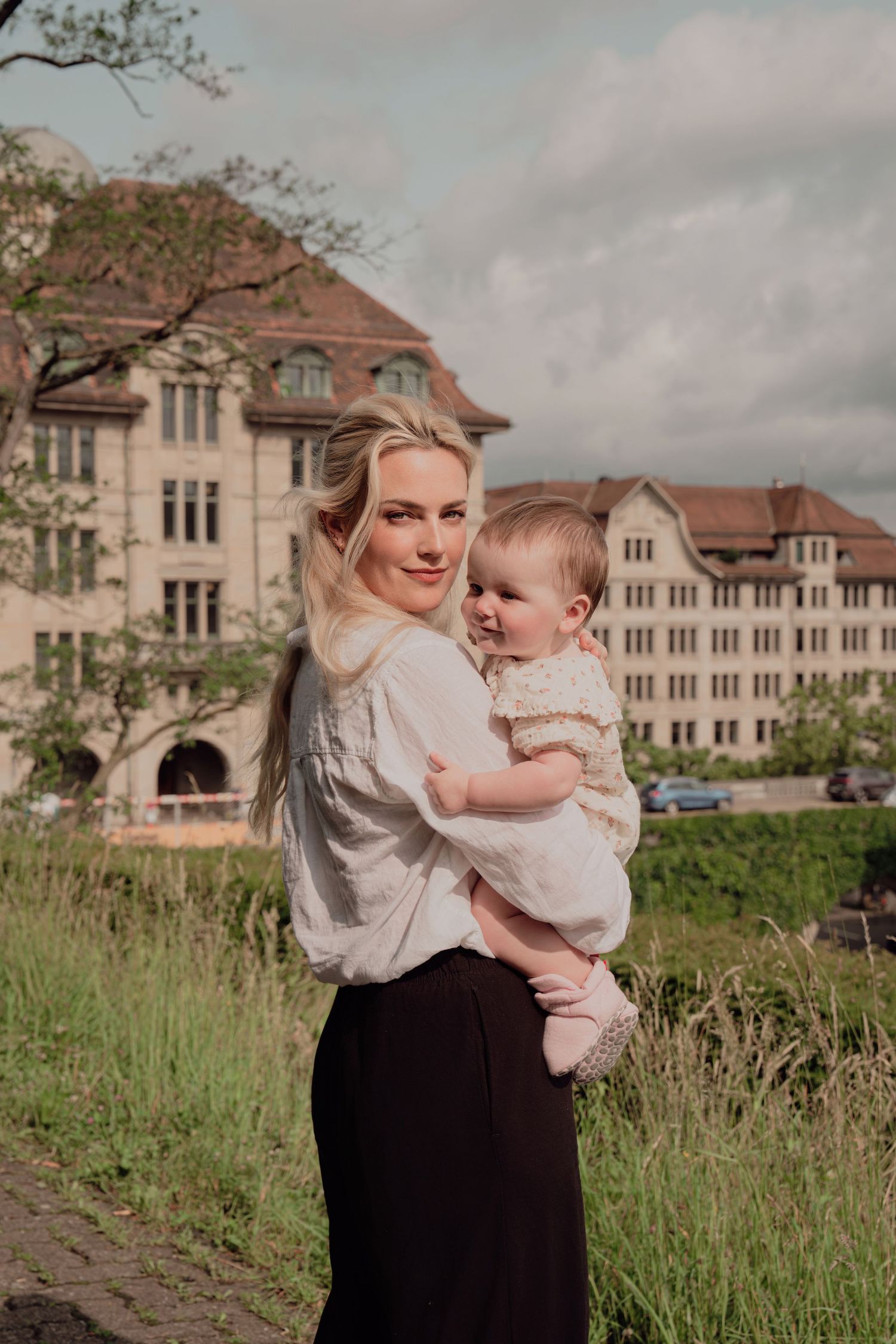 Blonde mother holding baby in arms, standing before historic European building on cloudy day.