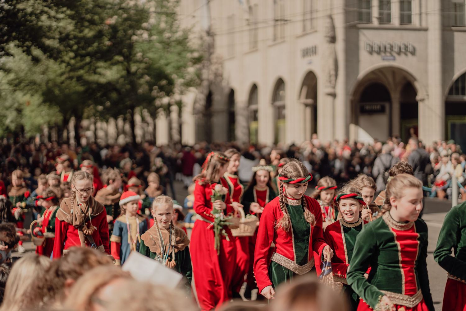 Children dressed in red and green historical costumes march in formation during a festive city street parade.