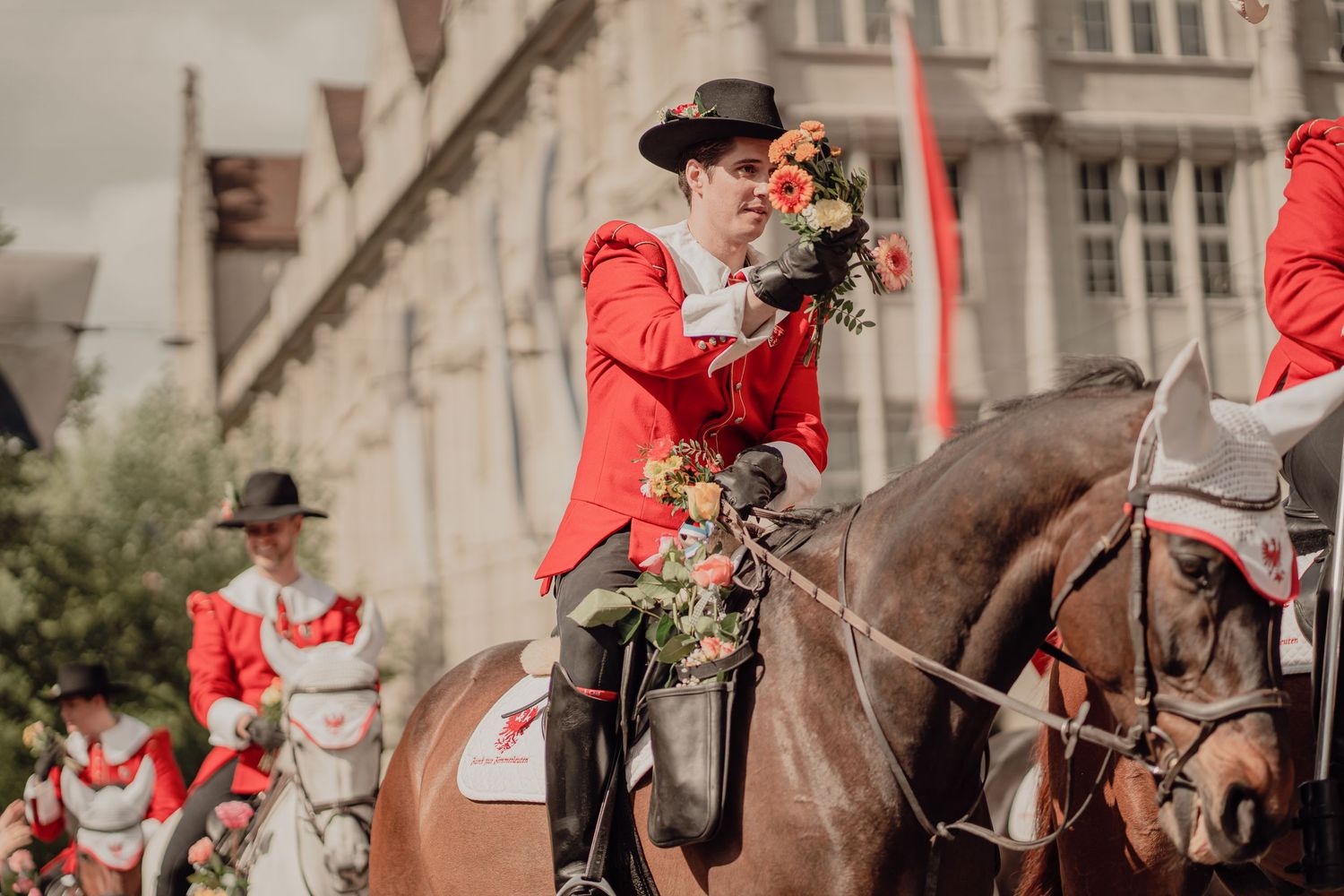 A rider in a red jacket on horseback holds a bouquet of flowers during a traditional parade.