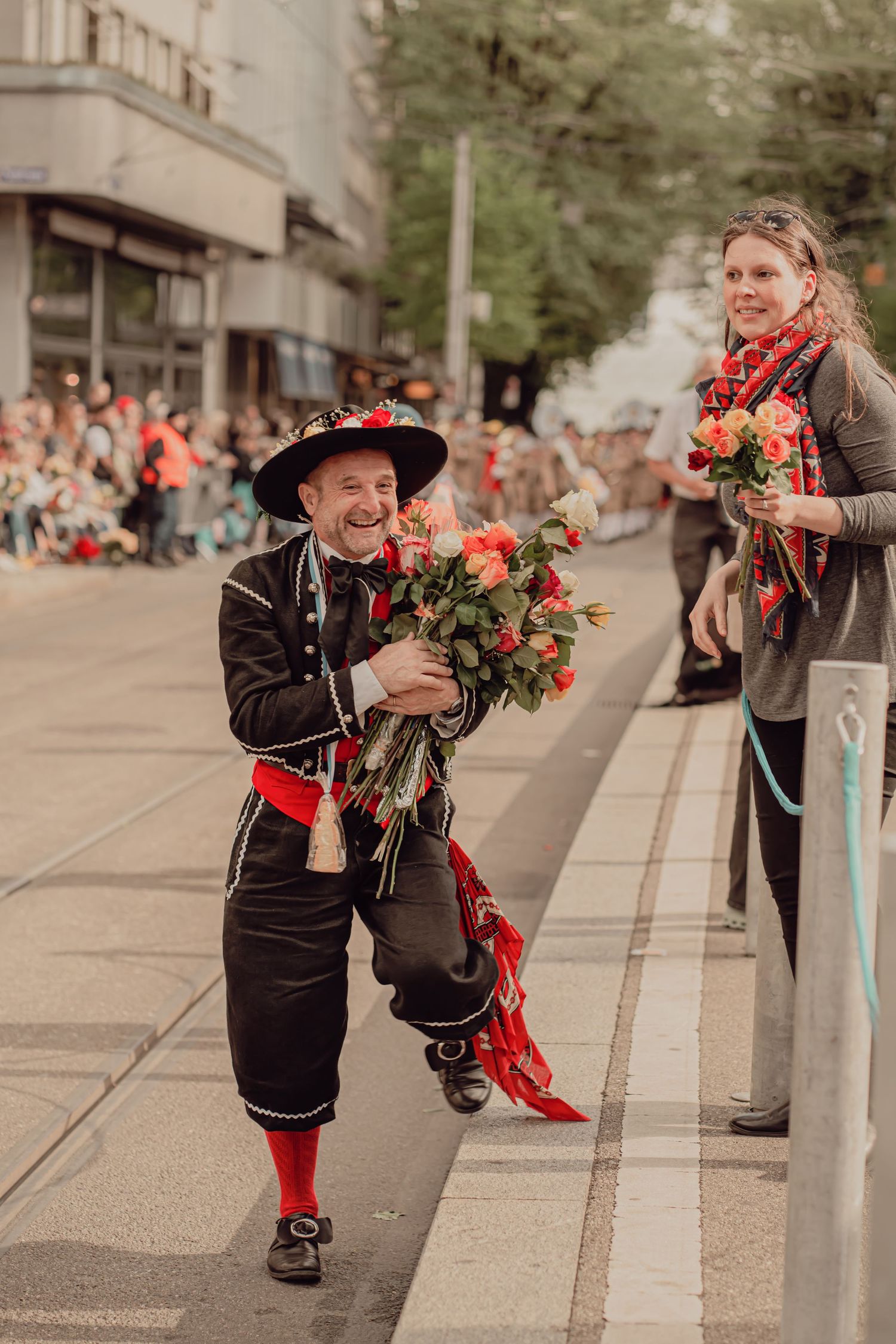 Cheerful man in traditional Swiss folk costume holds a large bouquet of roses at a street parade.