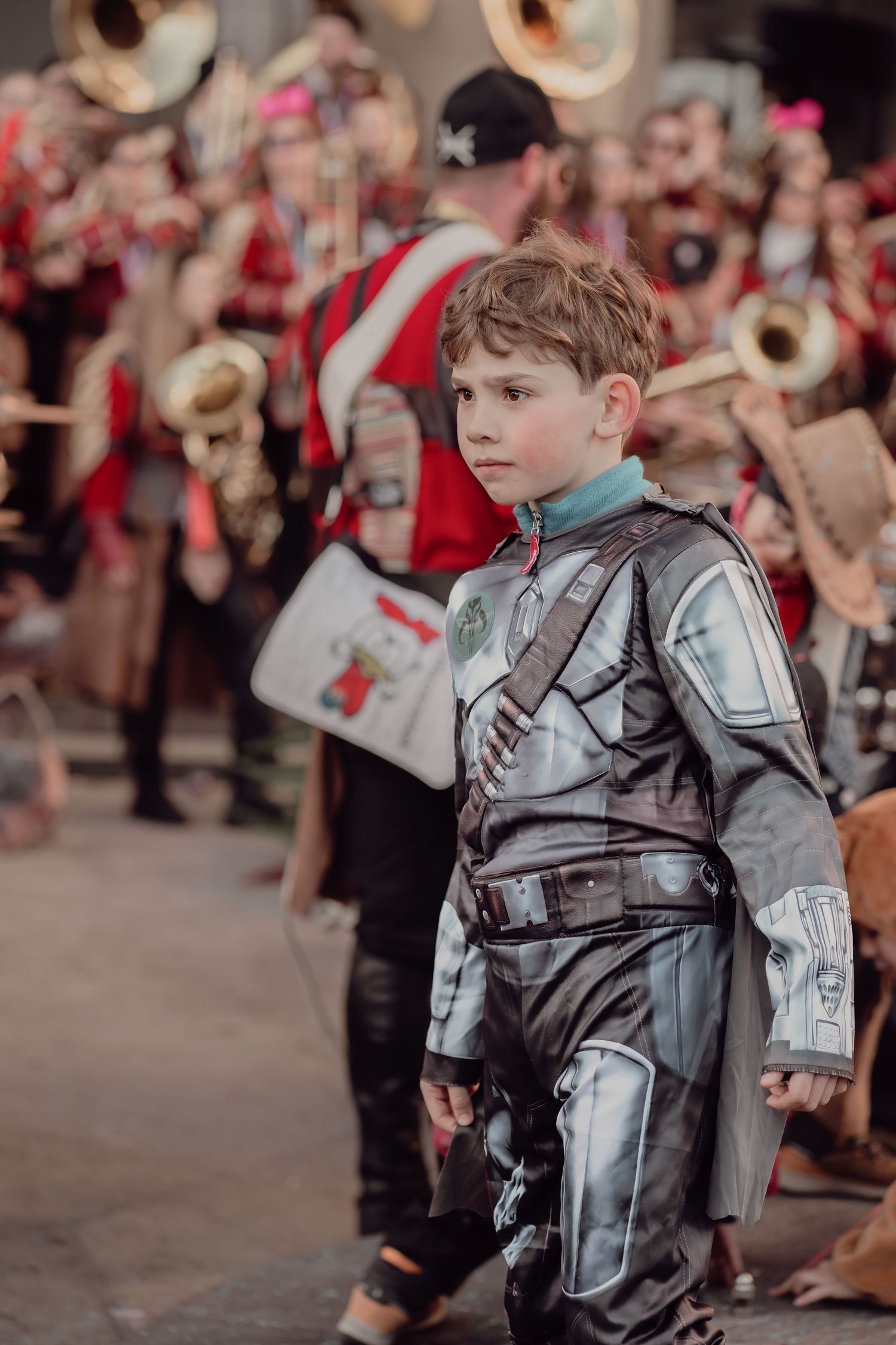 A boy in a silver Mandalorian-style armor costume walks in front of a marching band.