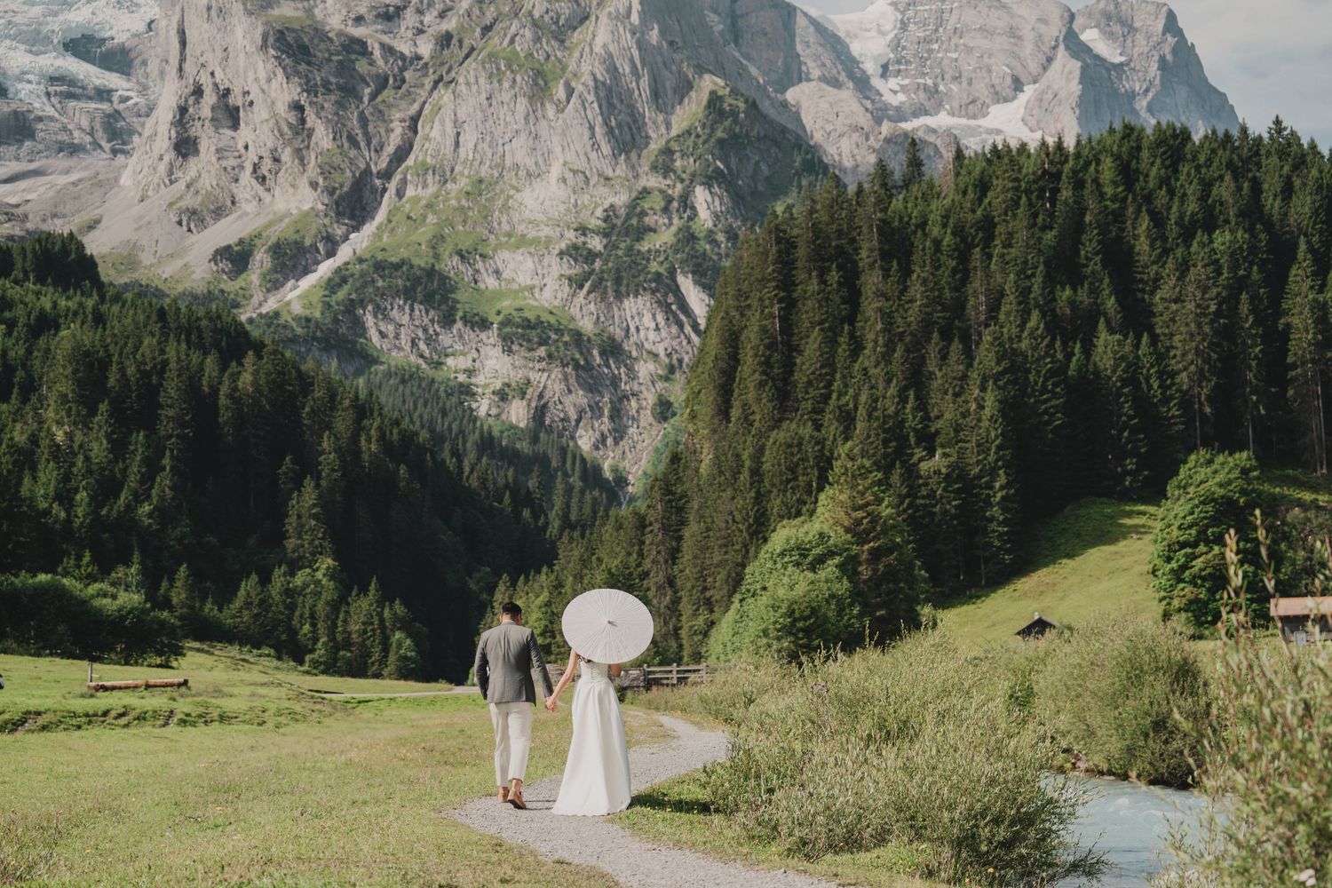 Newlyweds holding hands on a mountain valley path with white parasol, surrounded by lush green Alpine scenery.