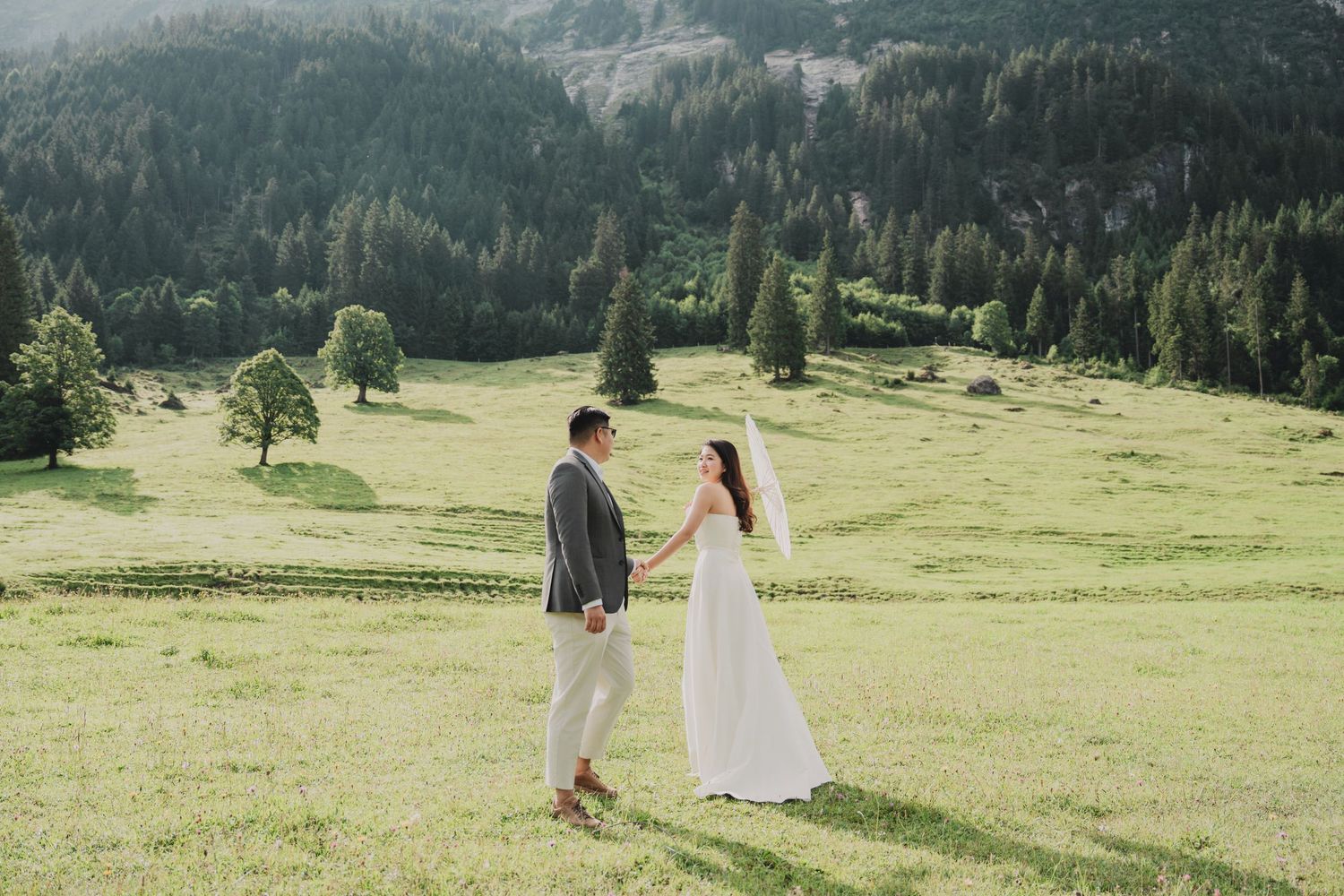Bride and groom holding hands in a lush green alpine meadow surrounded by pine forests during their wedding shoot.