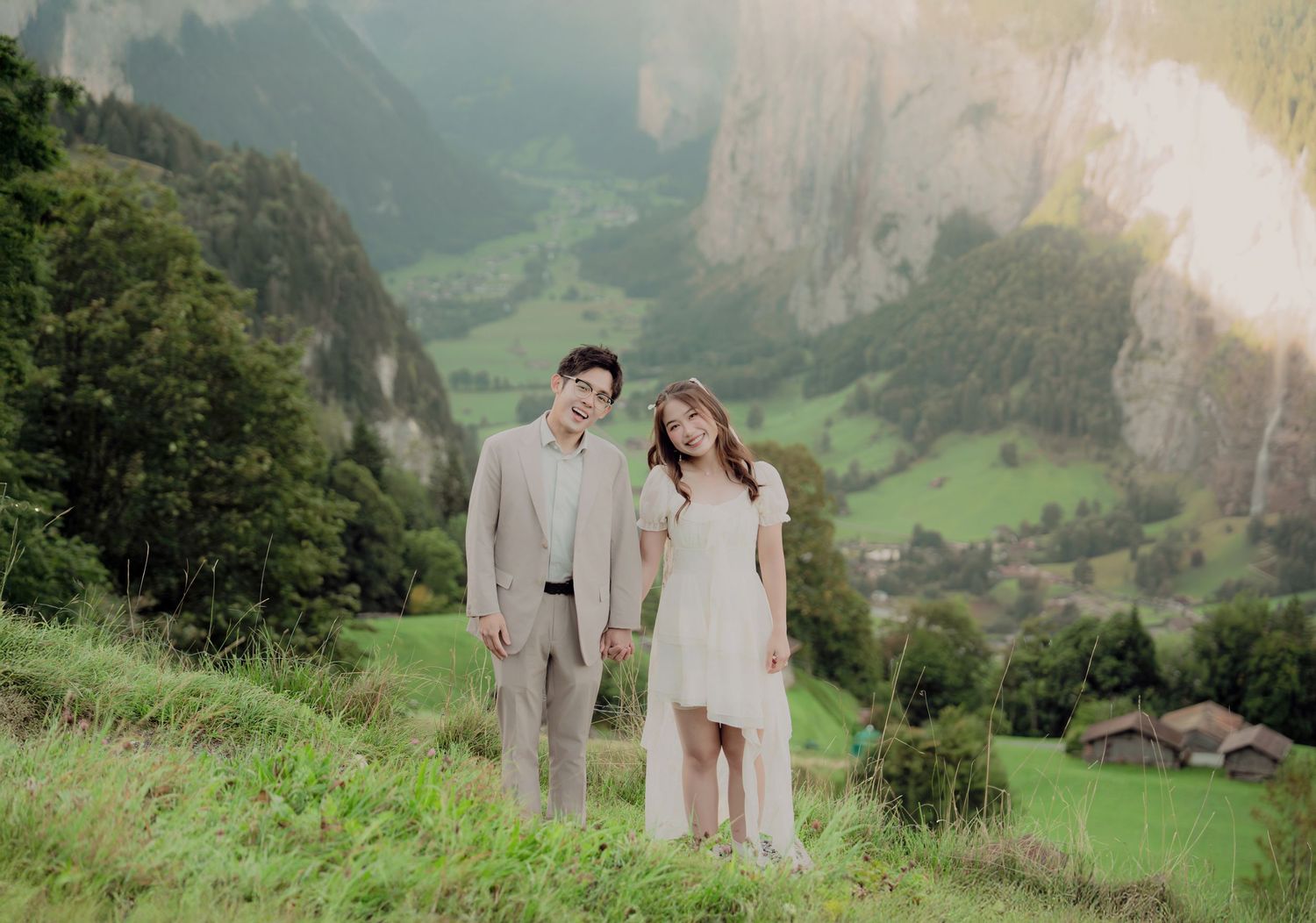 Couple in wedding attire holding hands on a green Swiss hillside with a stunning Alpine valley in the background.