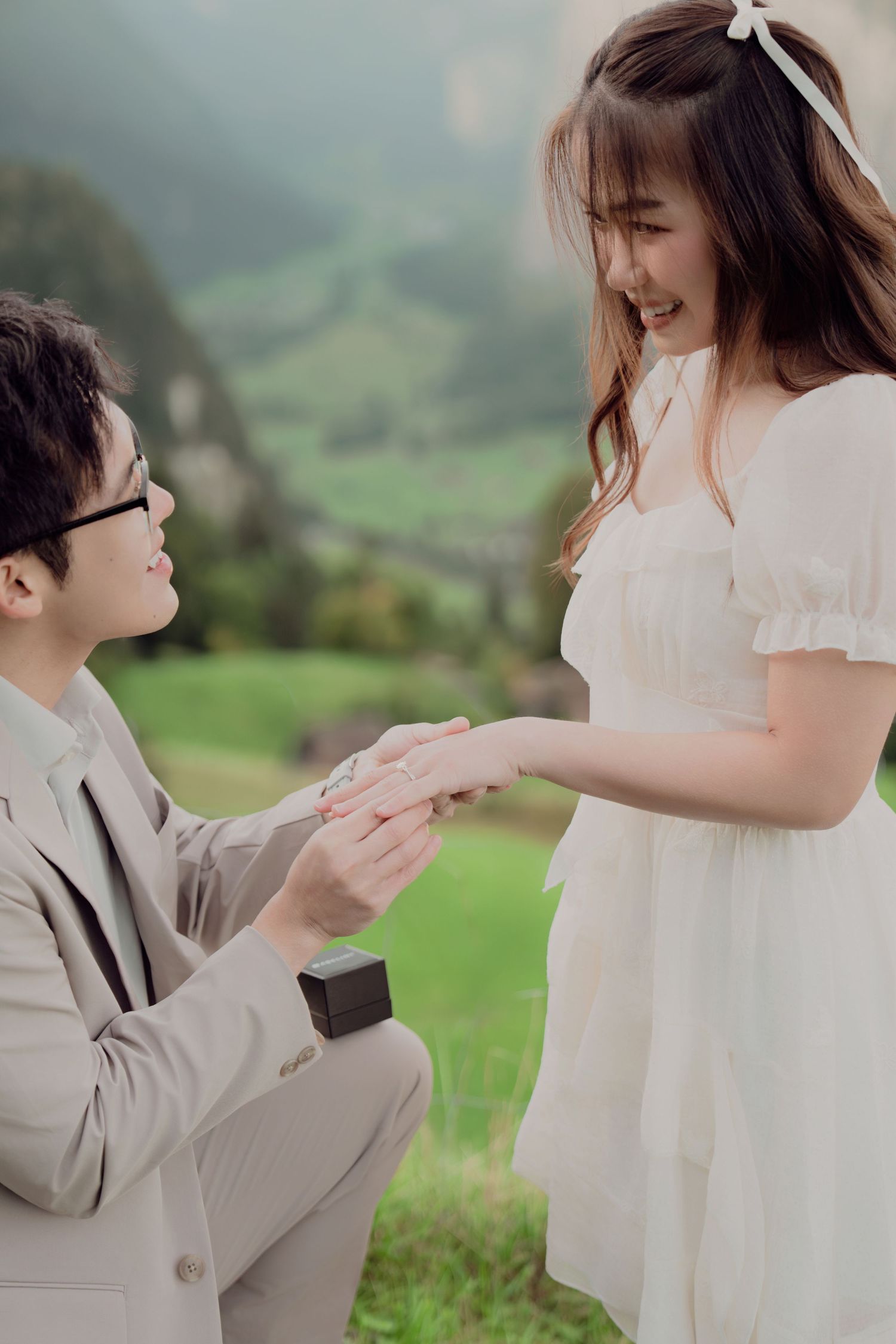 Man on one knee proposing with a ring to a smiling woman in a white dress in a mountain setting.