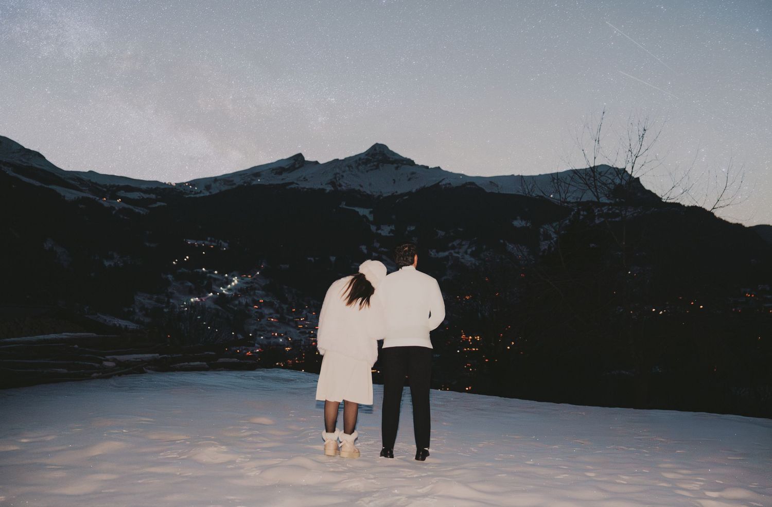 Couple standing together on snowy mountain landscape at dusk with city lights below.
