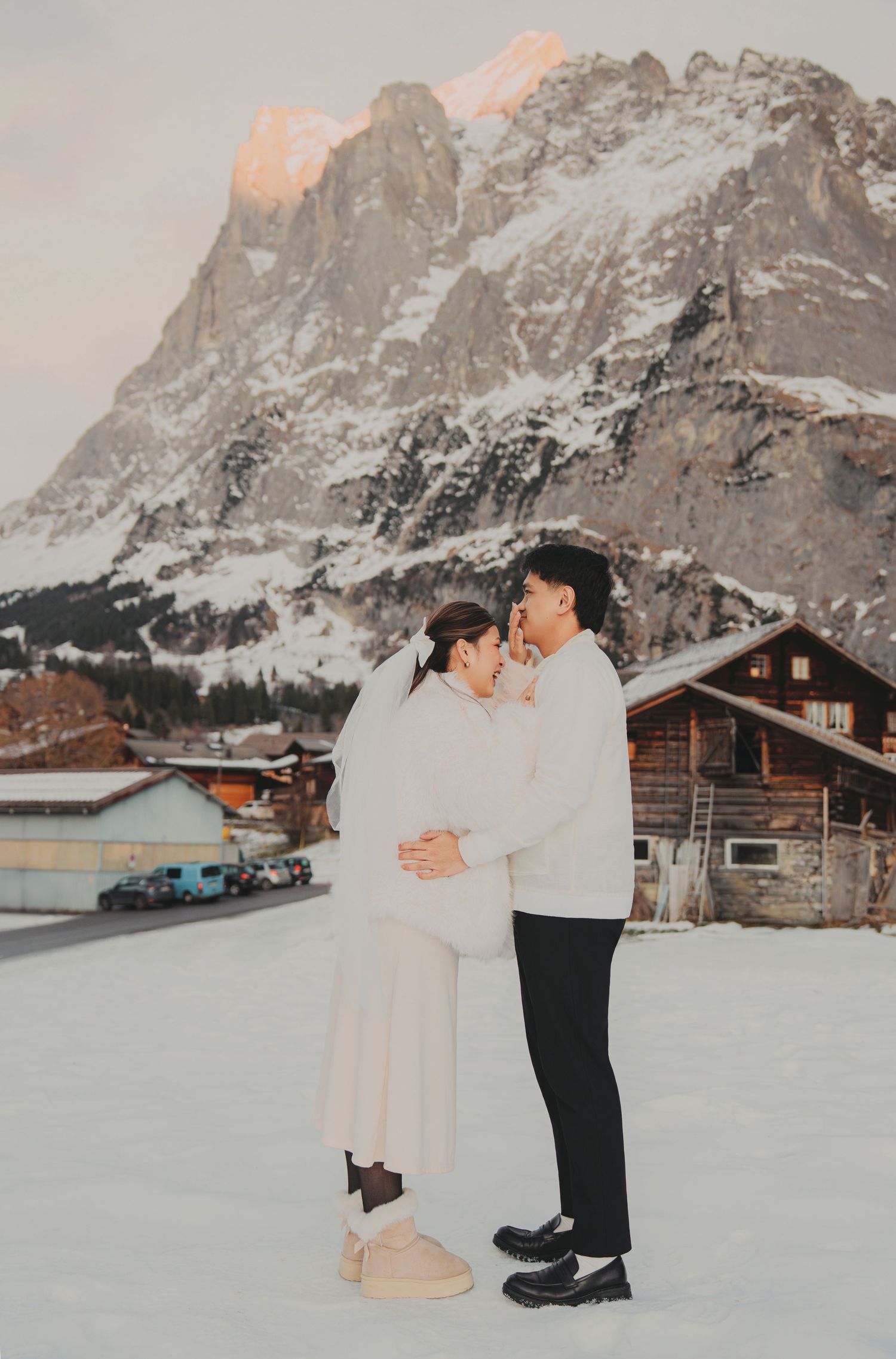 Romantic couple embracing in white outfits with a dramatic snow-covered mountain peak behind them.