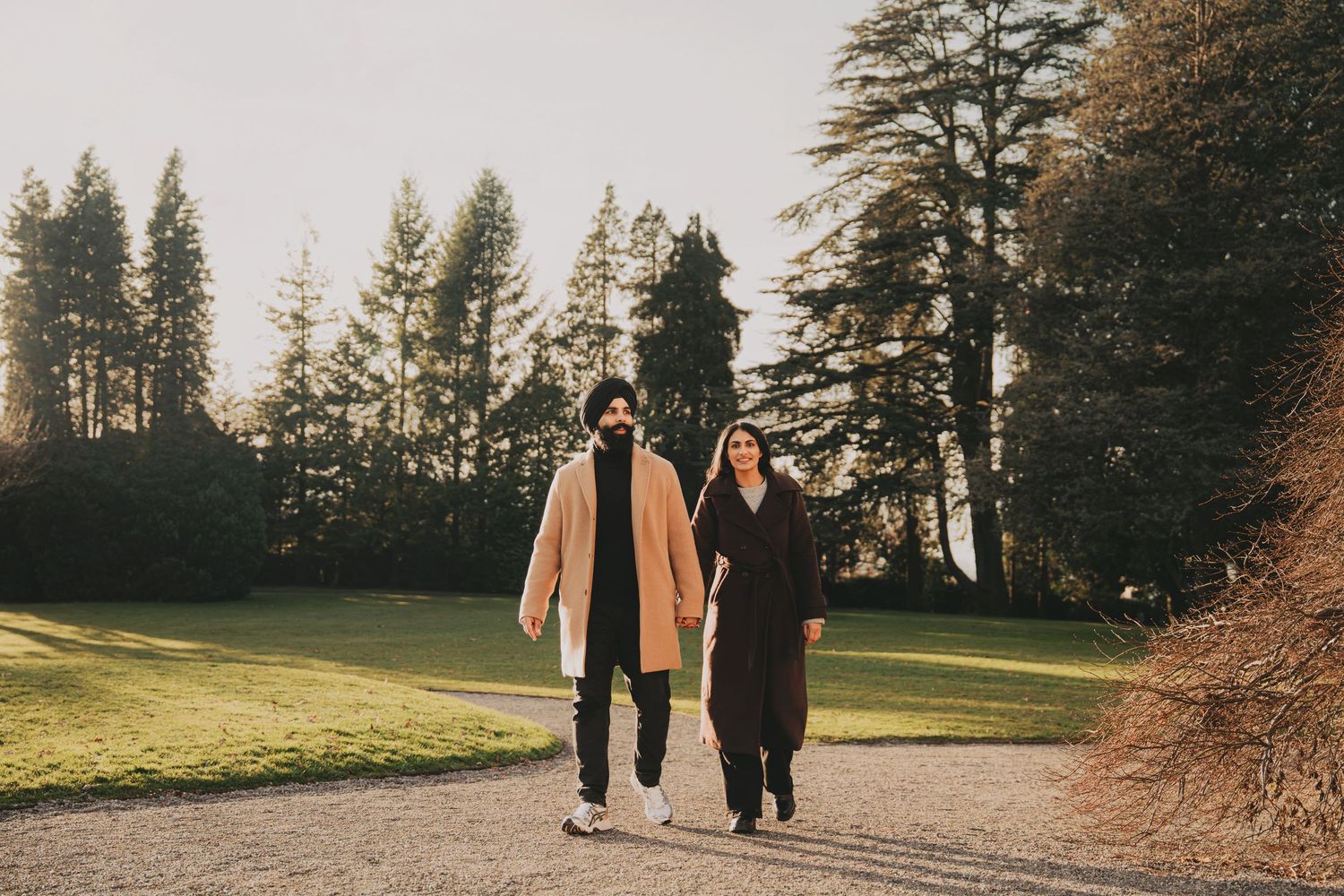 Man in beige coat and woman in black coat walking together on a scenic park pathway.