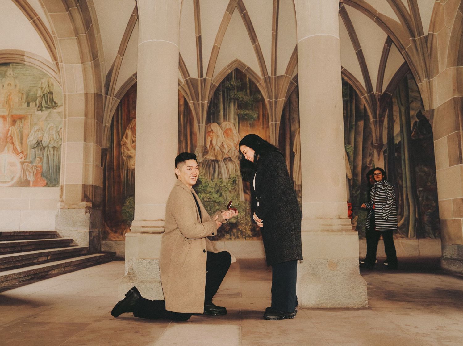 Man proposing on one knee inside gothic vaulted hall with medieval frescoes and onlookers.