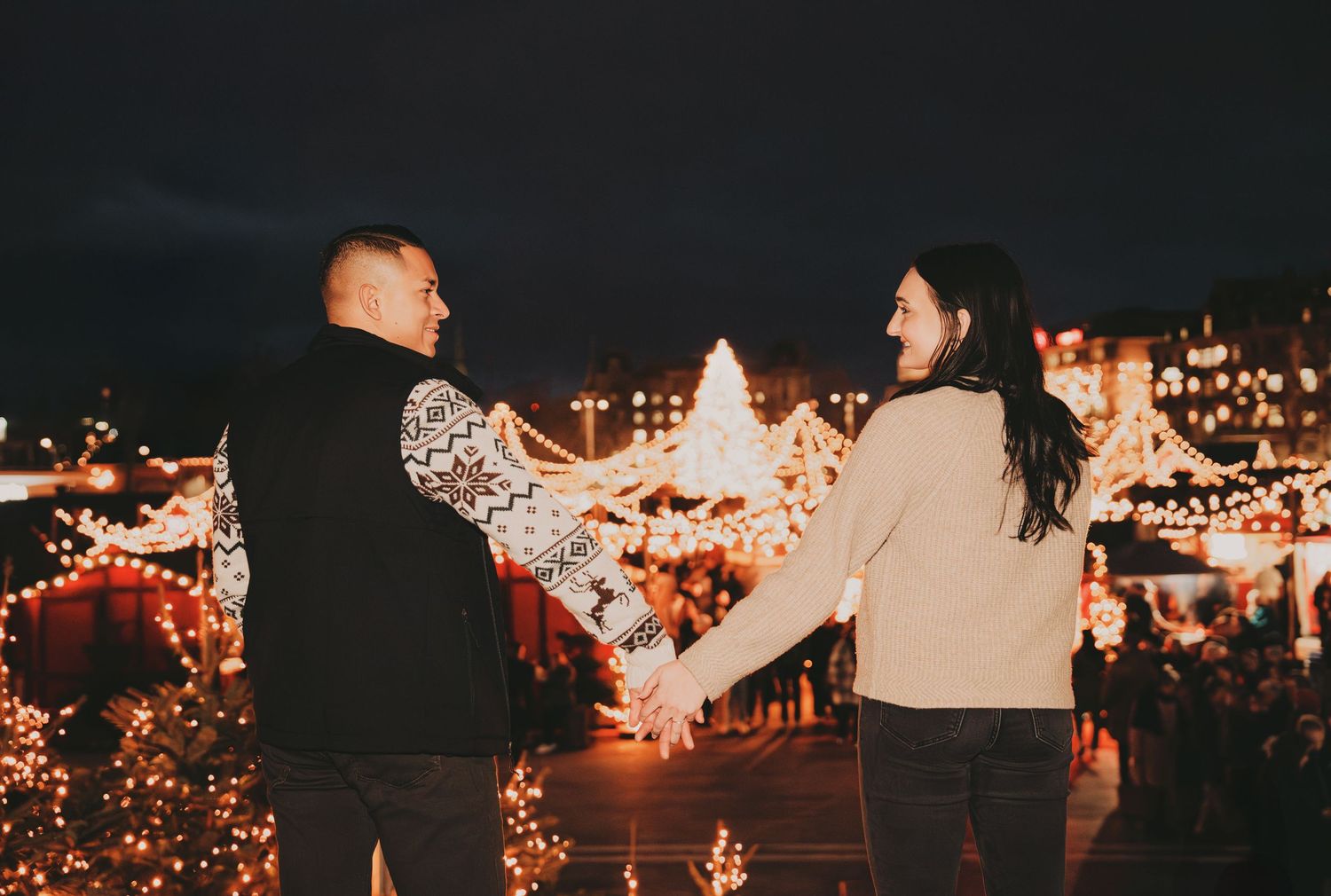 Couple holding hands and spinning joyfully at a festive Christmas market with a lit holiday tree.