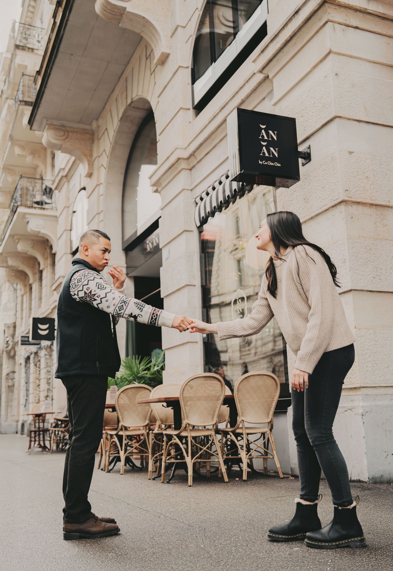 Couple holding hands outside AN AN restaurant with wicker chairs in a European city.